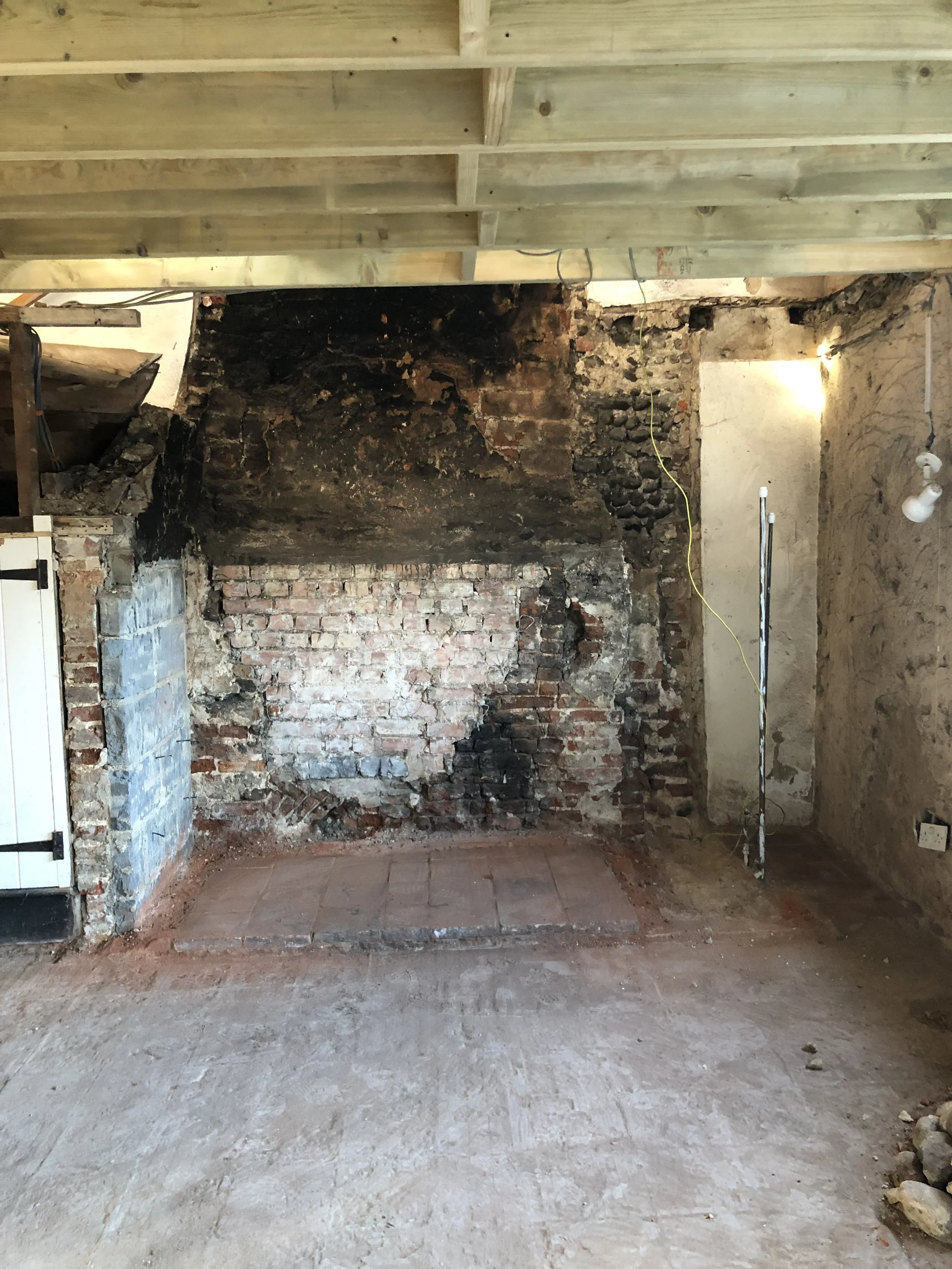 Interior of an unfinished room with exposed brick and charred blackened wall, partly covered with new wooden flooring and includes construction wiring and tools.