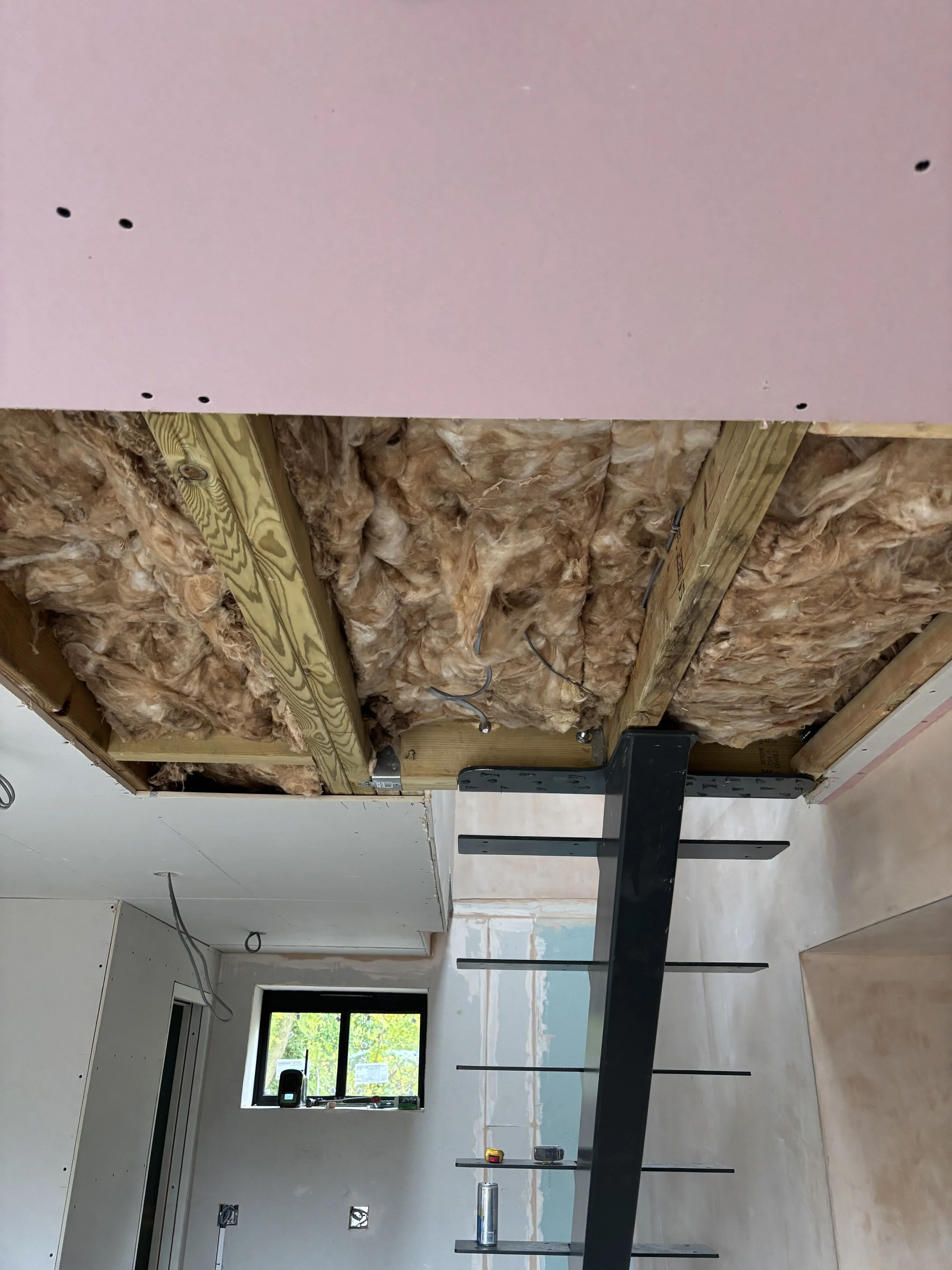 An unfinished room with insulation and wooden framing in the ceiling area, a black metal railing in the foreground, a window with green trees outside, and construction tools and wiring visible.