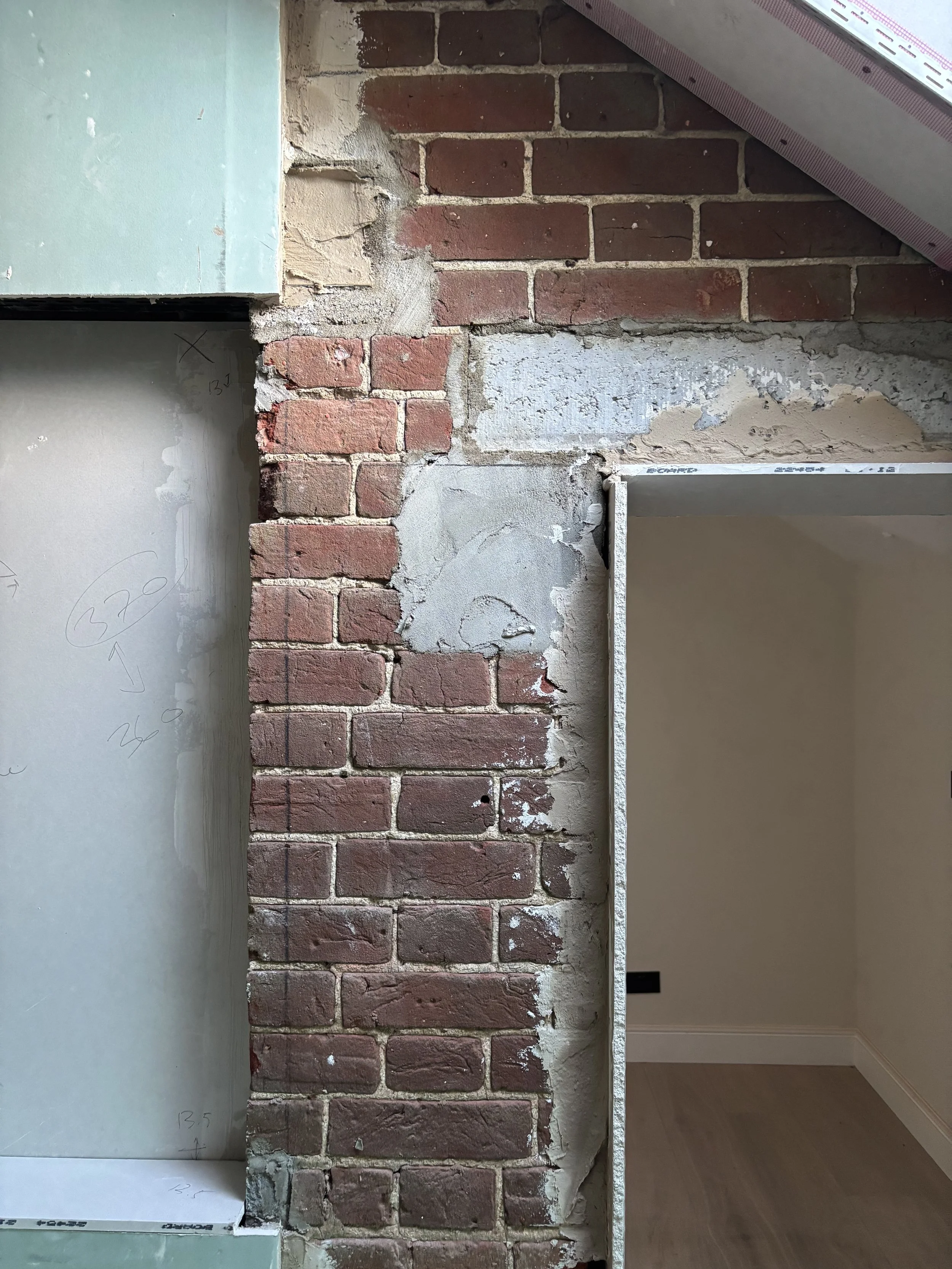 Interior wall showing exposed red brick with some mortar and concrete damage, adjacent to a green drywall with markings and a partially visible door frame.