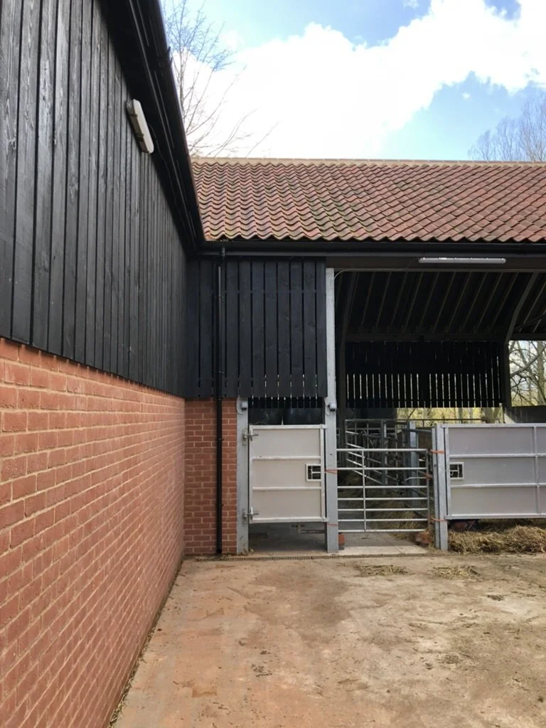 View of a farm or equestrian facility with a brick wall, metal gate, and black wooden building with a red-tile roof, under a partly cloudy sky.