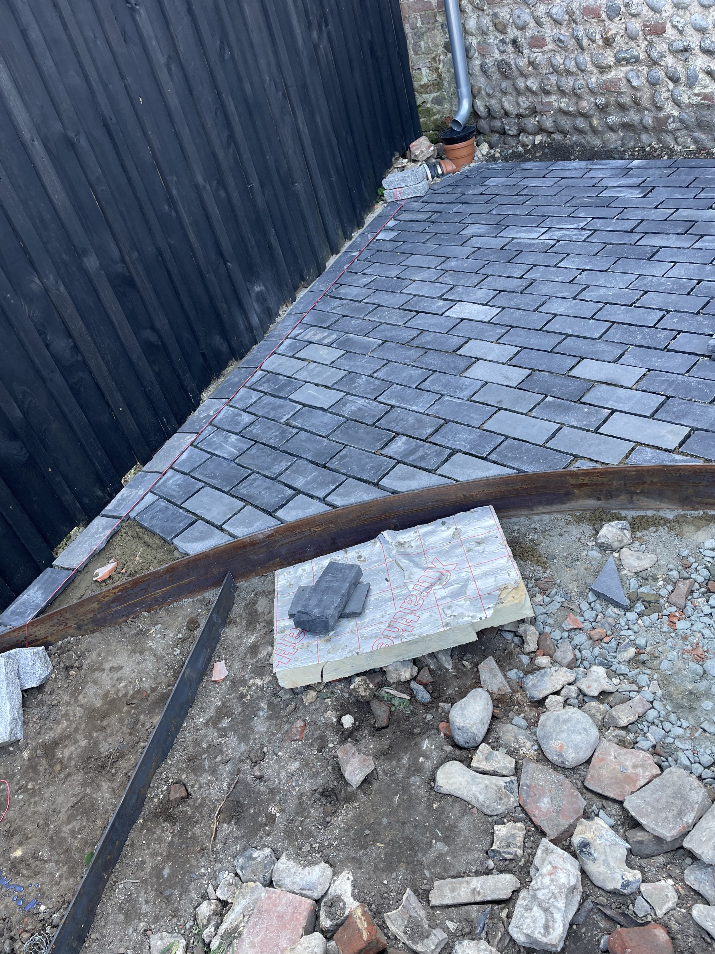 Construction site with cobblestone pavement, black wooden fence, and stone wall. Construction materials include concrete blocks, bricks, and metal support beams. A red string line marks the edge of the paving area, and a drainpipe is attached to the 