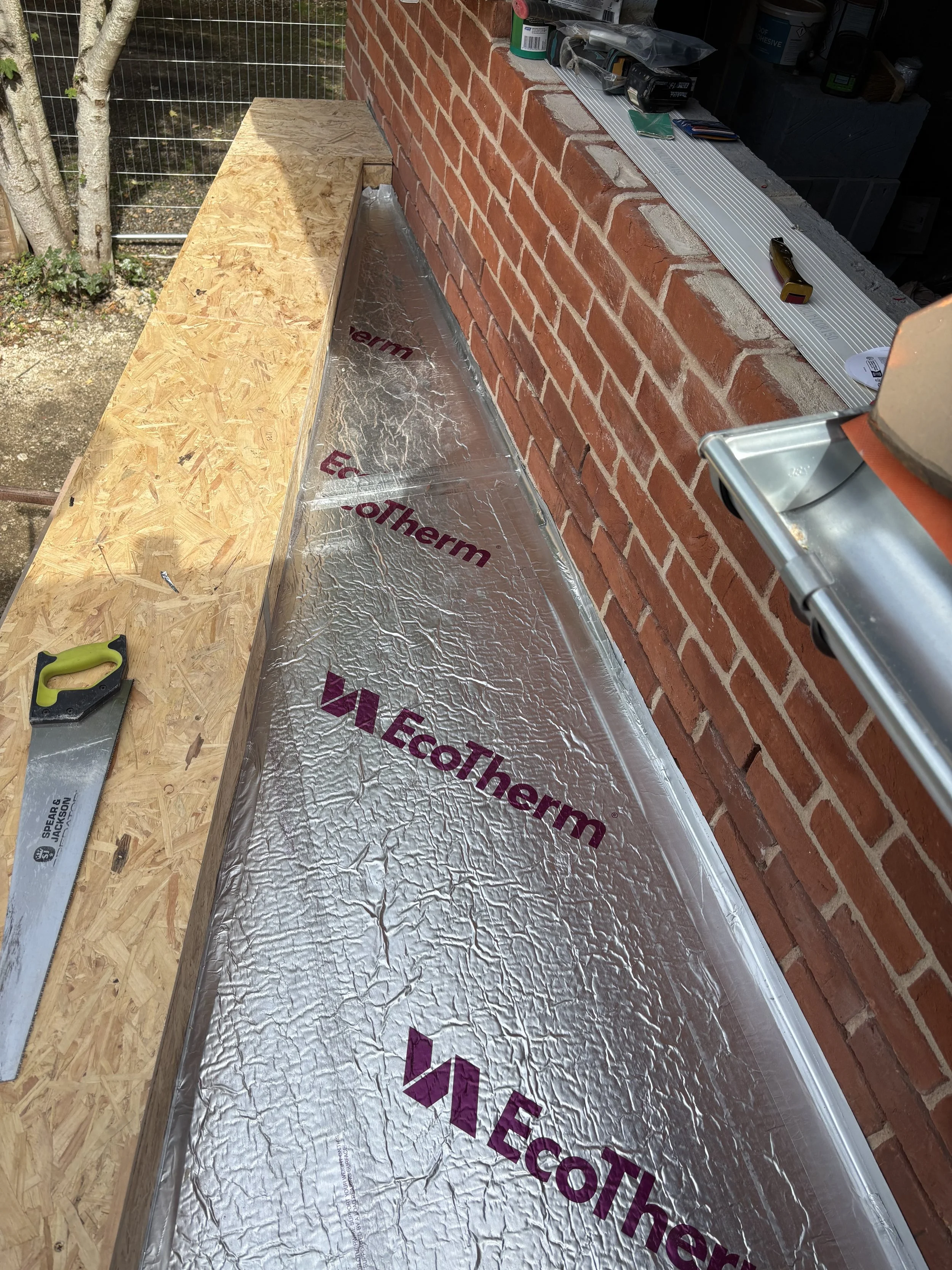 Construction scene showing a wooden platform, a brick wall, and construction tools such as a saw, measuring tape, and screwdriver. Insulation material labeled 'EcoTherm' is also visible.