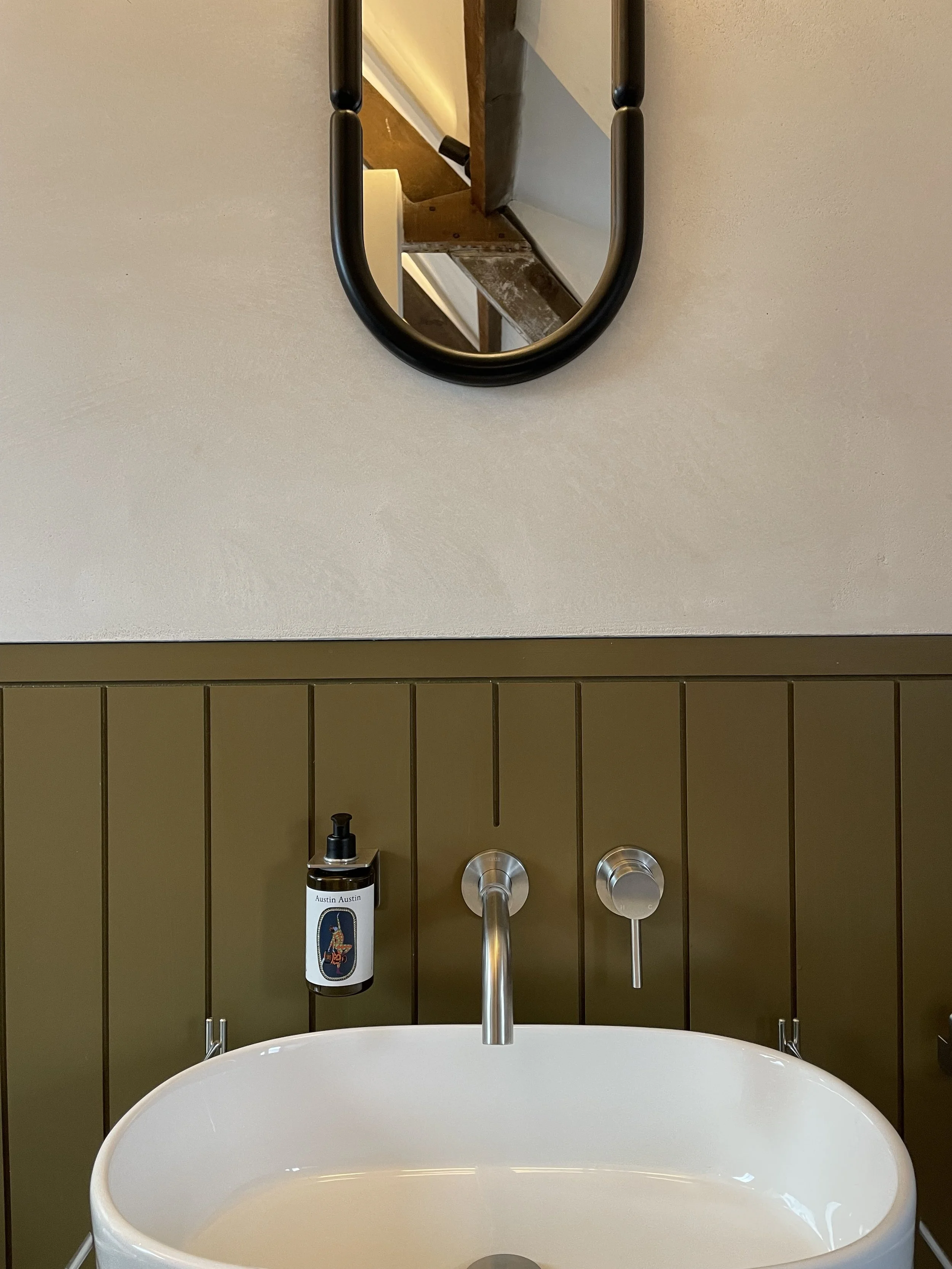 Close-up of a bathroom sink with a silver faucet and two silver knobs, a soap dispenser labeled 'Austin Austin,' mounted on a dark brown tiled wall. Above the sink, a large mirror reflects the ceiling and parts of the wall.