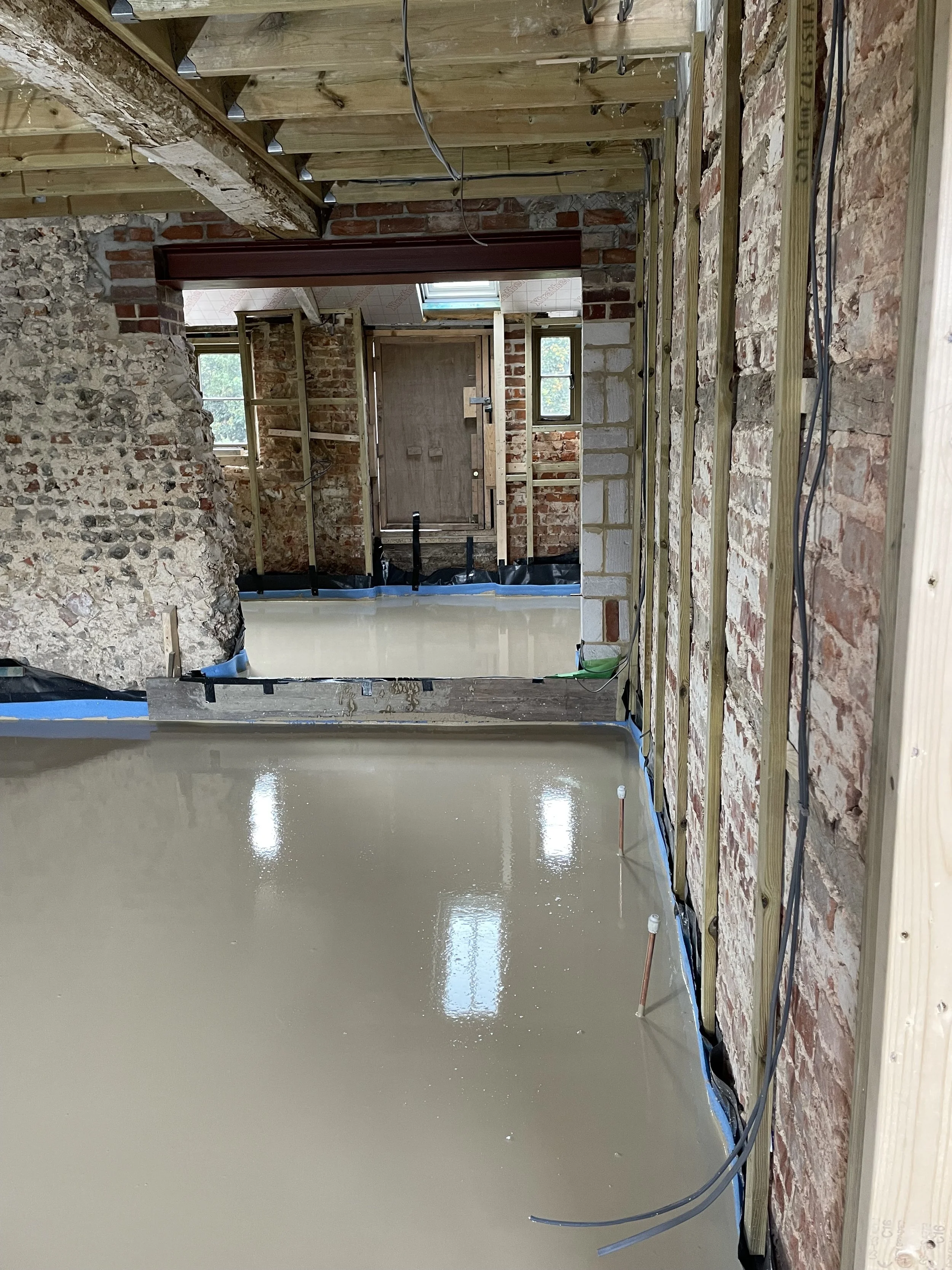 Interior of a house under renovation showing a newly poured concrete floor, exposed brick and stone walls, wood framing, electrical wiring, and windows.
