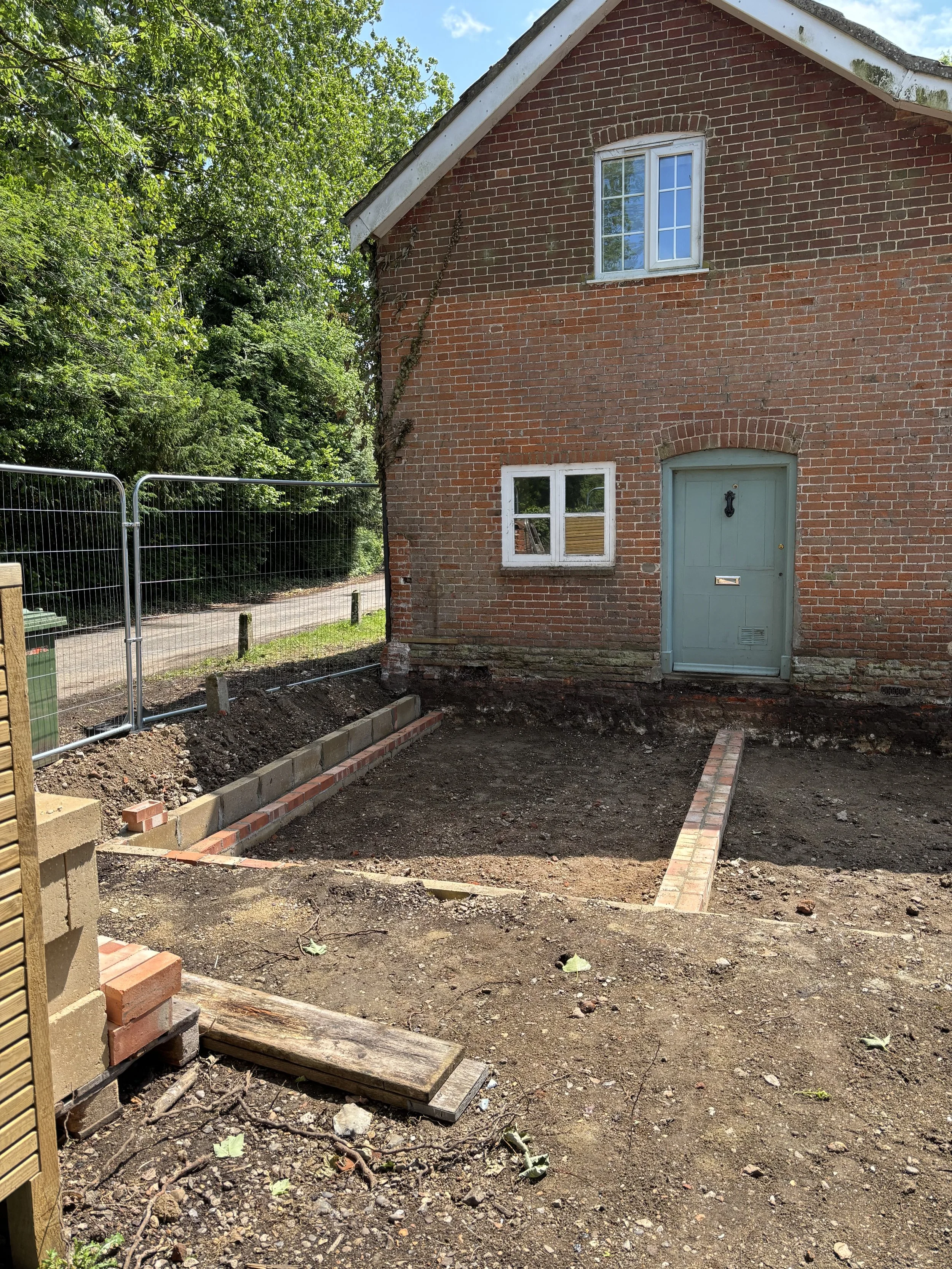A brick house with two windows and a door, partially under construction or renovation with exposed soil and bricks in the foreground, a green tree to the left, and a blue sky with some clouds above.