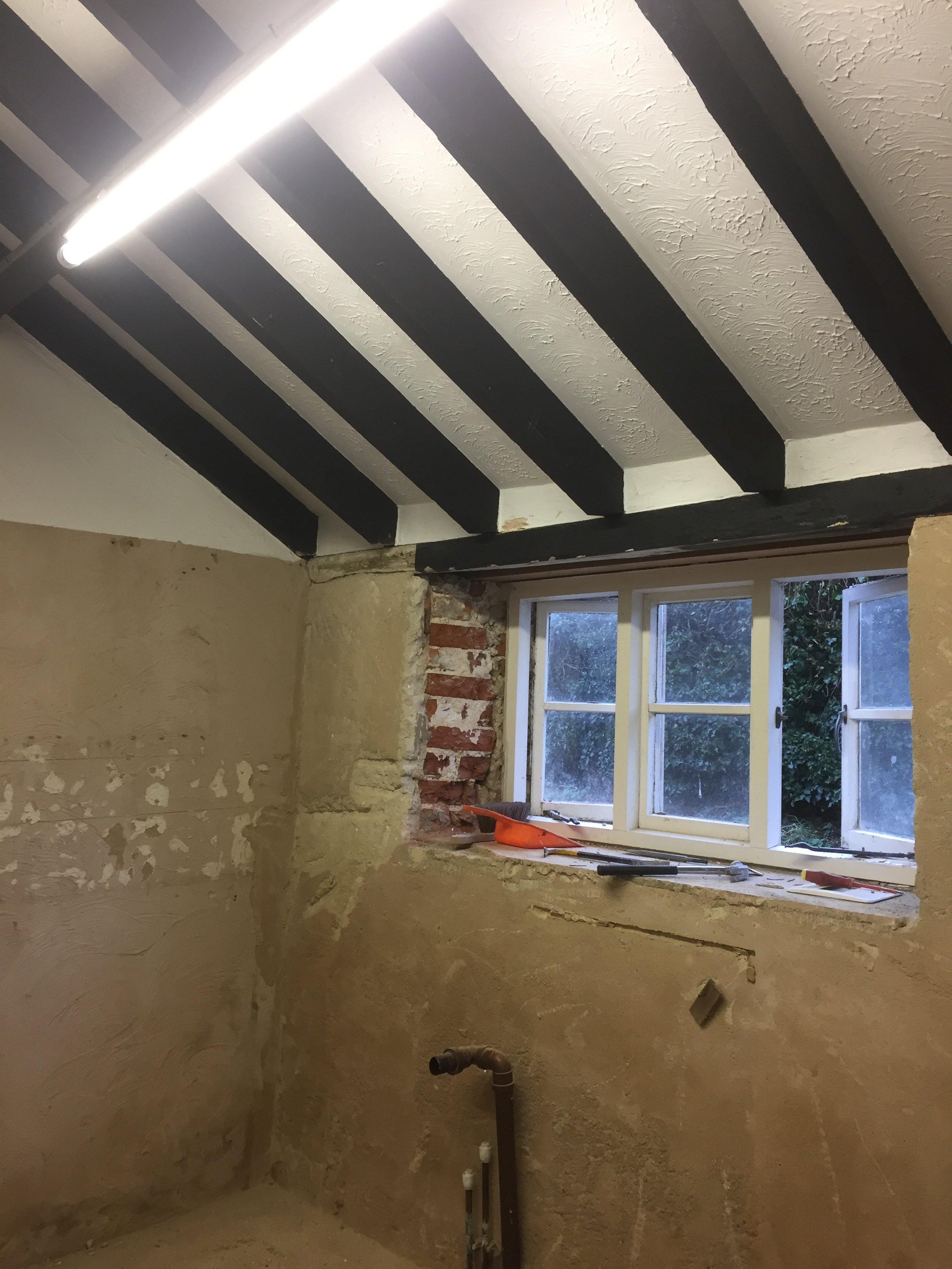 Partially renovated room with a window showing outdoor greenery. The ceiling has dark wooden beams, and construction tools and pipes are visible on the unfinished wall.