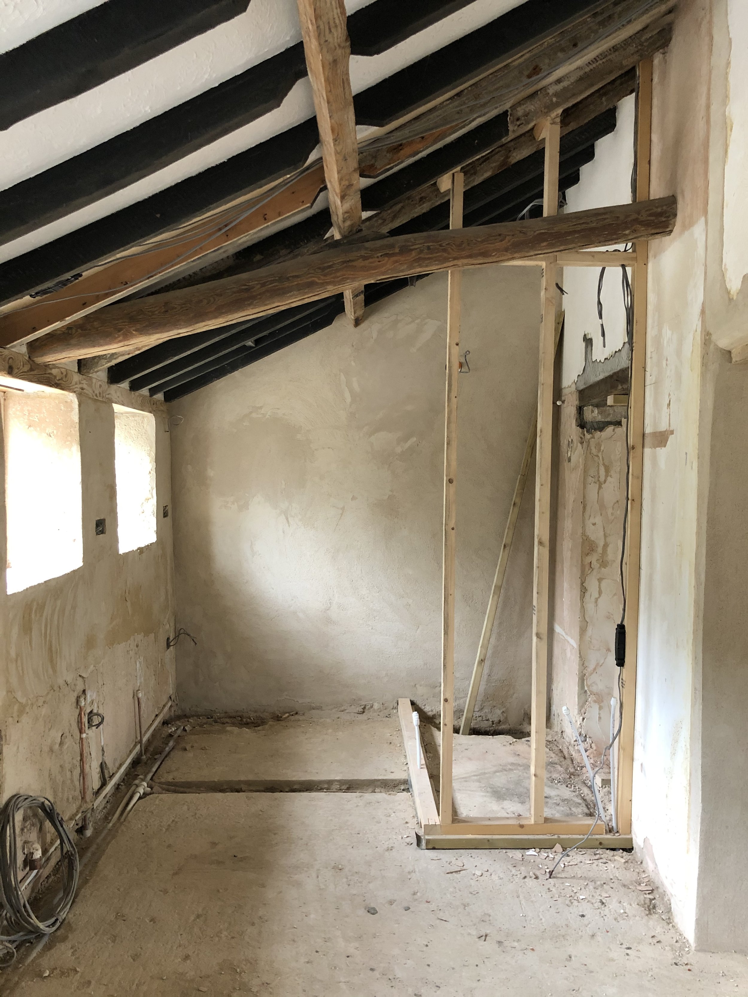 A room under construction with exposed wooden framing, black beams on the ceiling, and unfinished walls. There are electrical wires on the walls and cables on the floor.