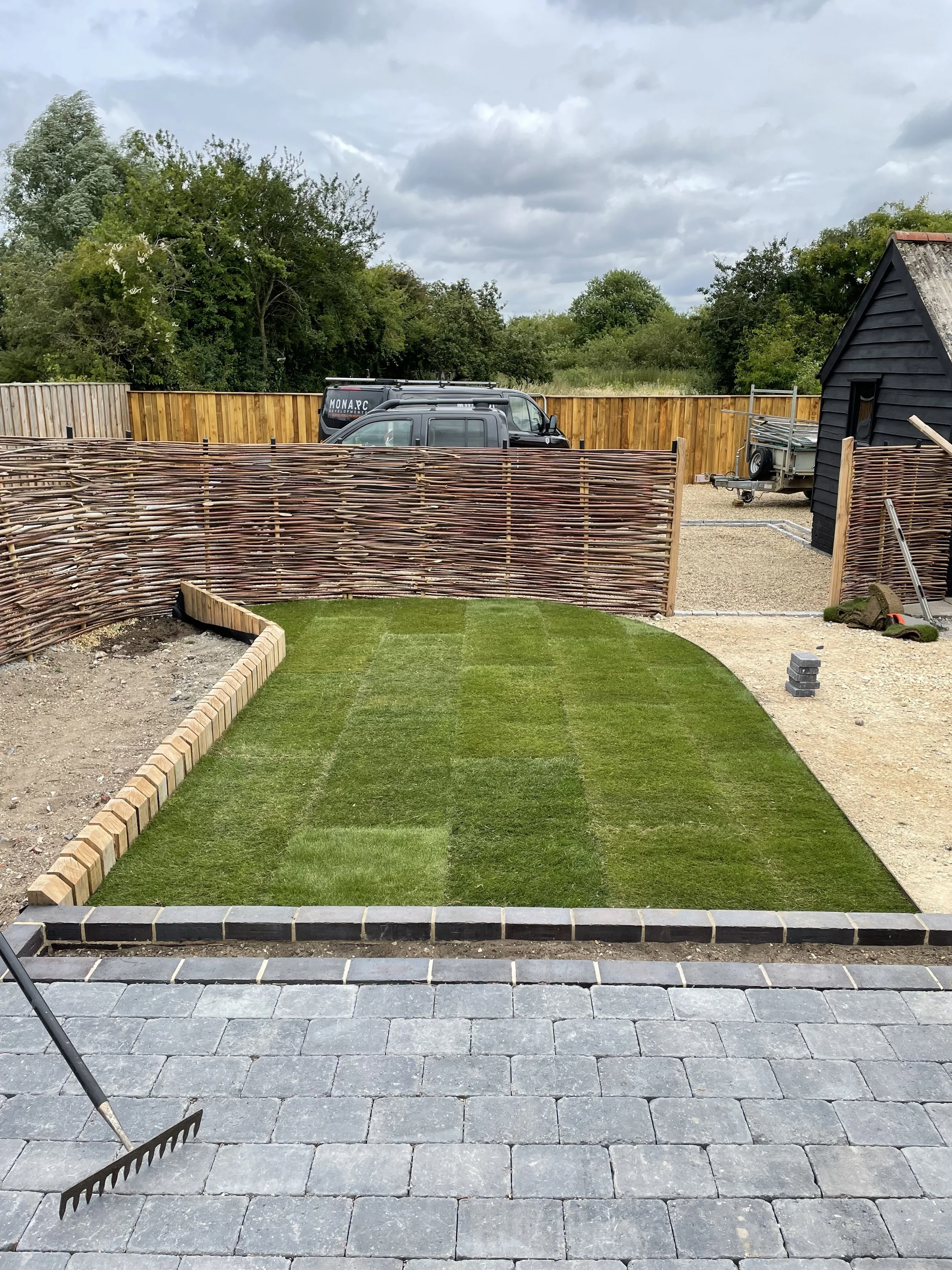 A backyard under construction with a newly laid paved patio, a green lawn, a wooden privacy fence, a small black building, construction tools, and vehicles parked in the background, under a cloudy sky.