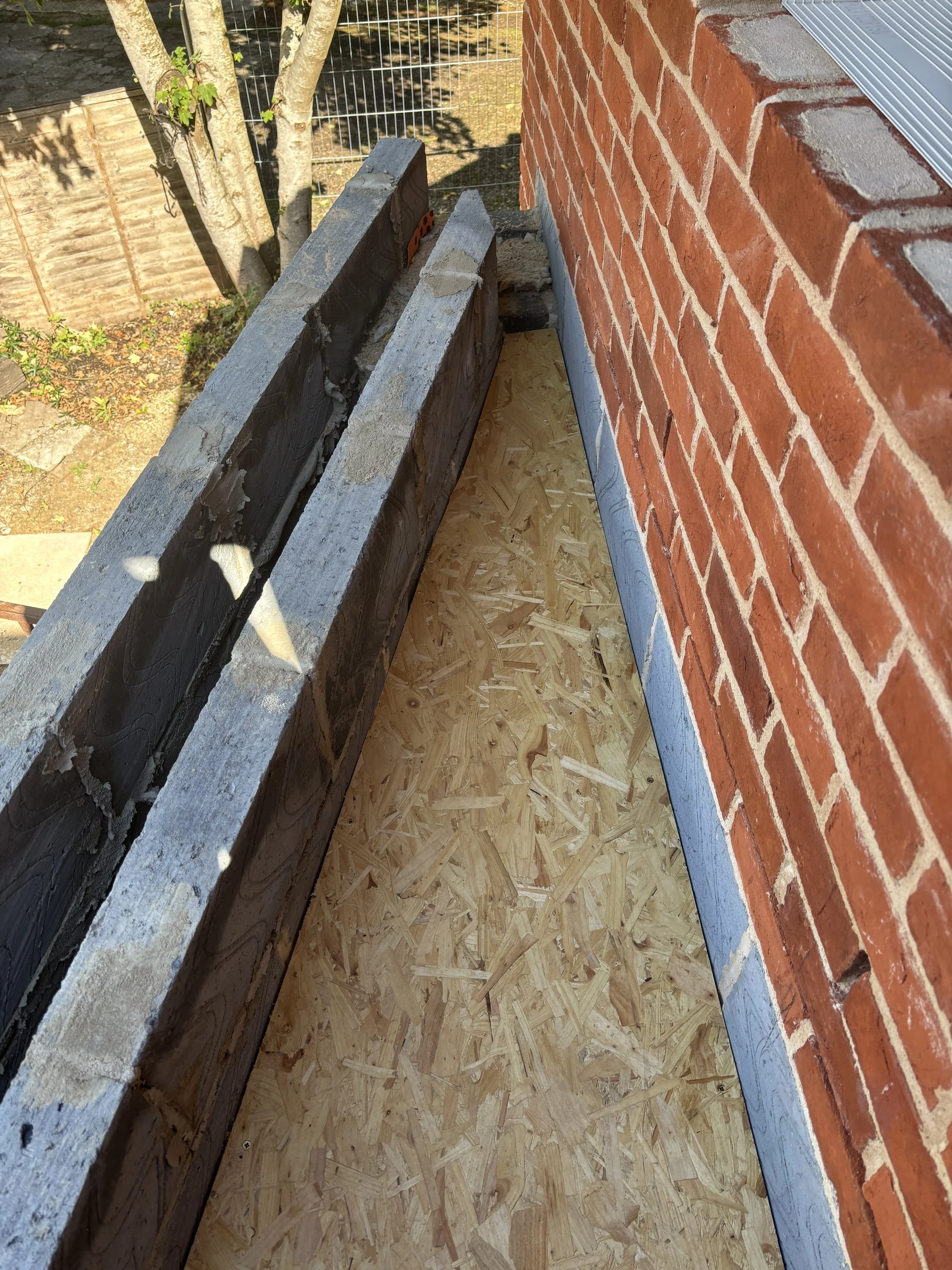 Close-up view of a construction site showing a brick wall, an under-construction wooden floor, and some wooden planks placed horizontally. A metal coping is visible on the brick wall's edge, and a small tree with green leaves is present in the backgr