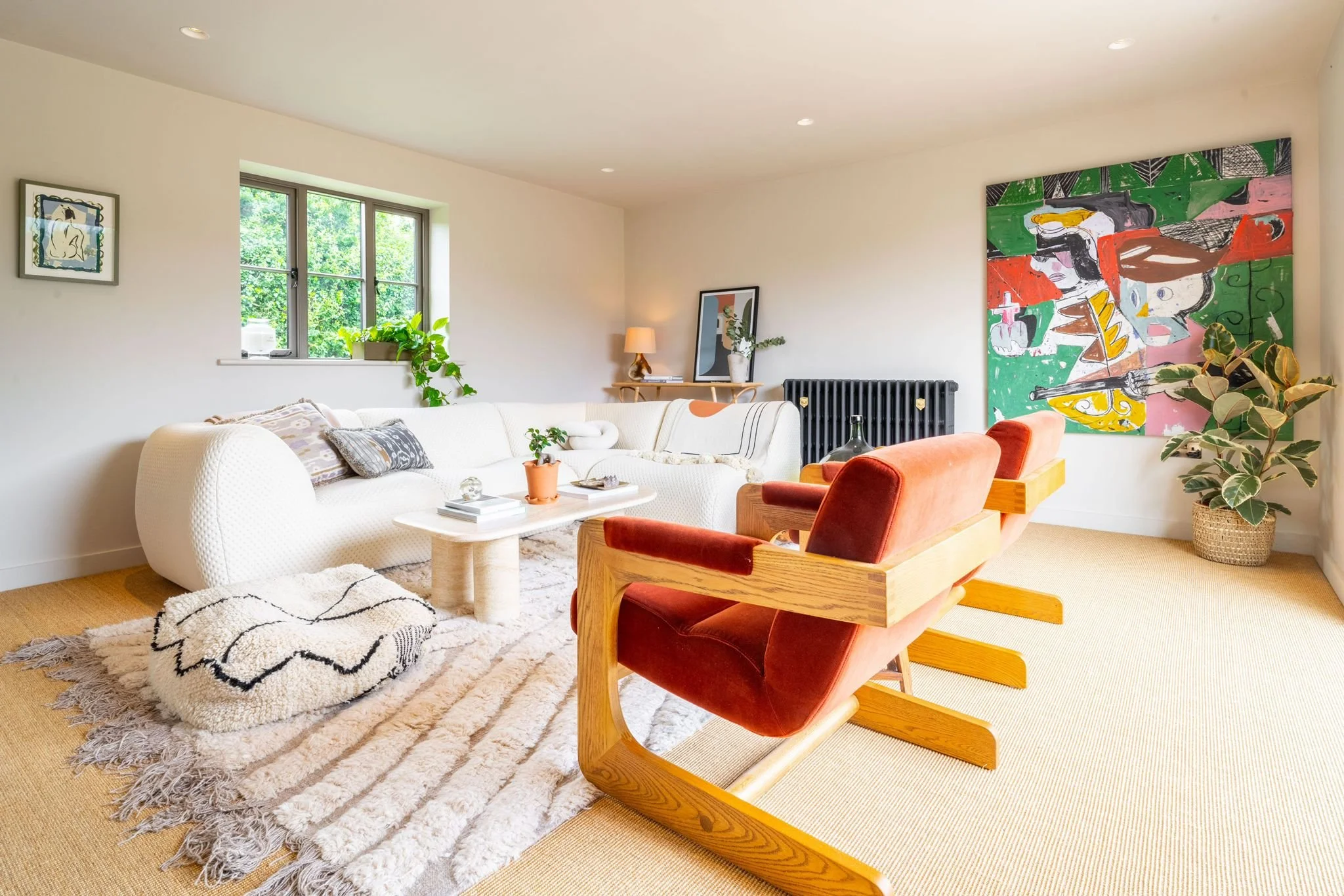 A living room with a white sofa, orange and red chairs, a wooden side table with a lamp and decor, a large colorful abstract painting, green houseplants, a window with greenery outside, a black radiator, and a textured beige rug.