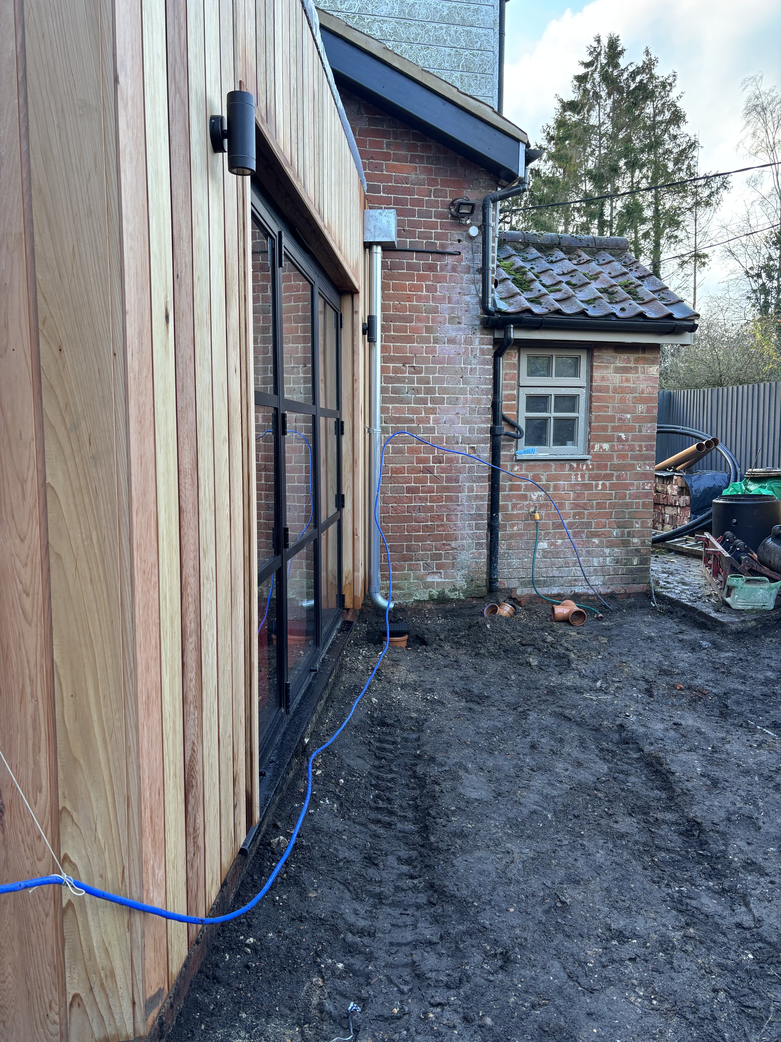 Backyard construction site with exposed soil, brick house with a small window, black drain pipes, and a wooden wall on the left side. Various construction tools and materials are visible.