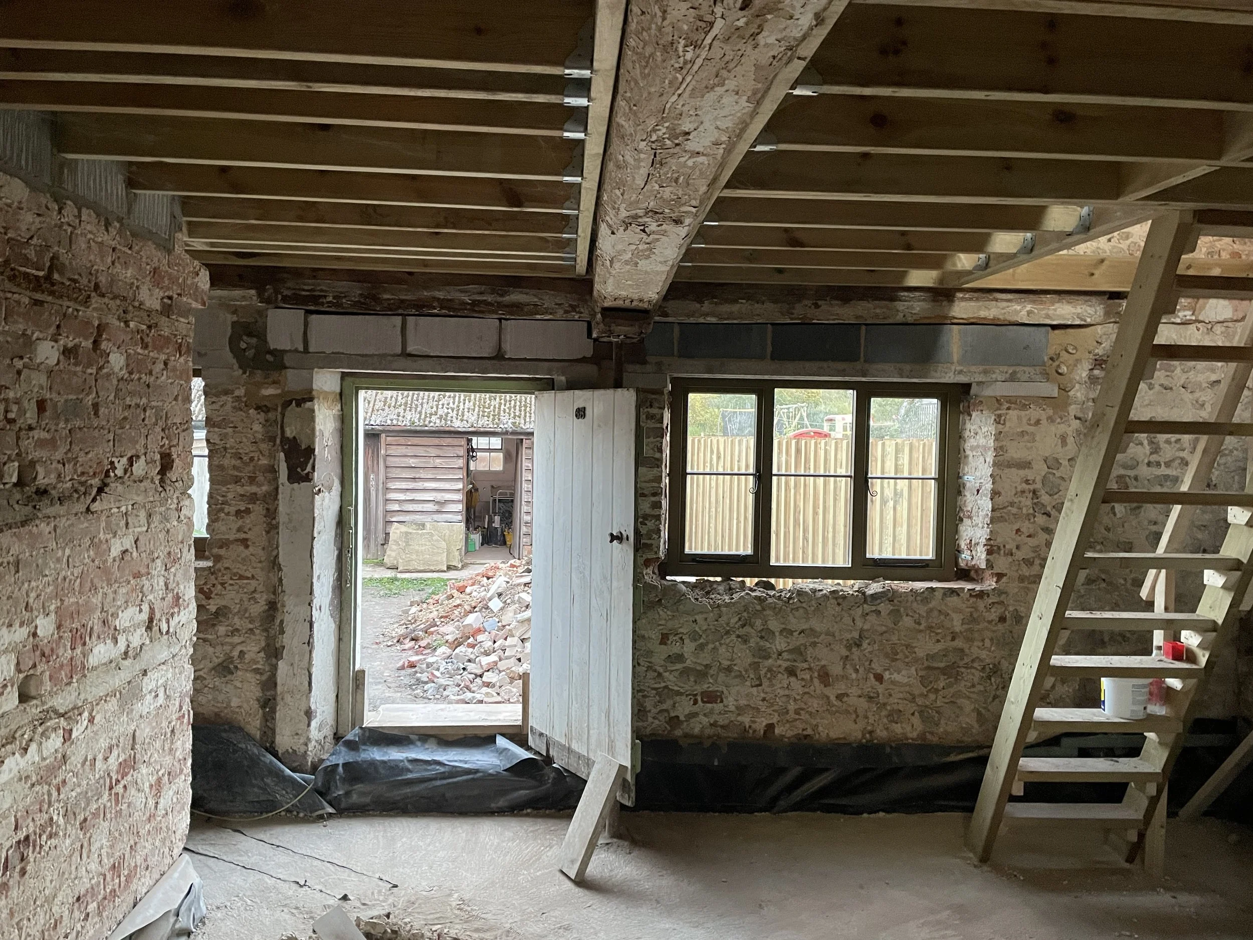 Interior of a building under construction with brick walls, wooden framing, and an open doorway leading outside to a yard with a wooden fence and a shed.