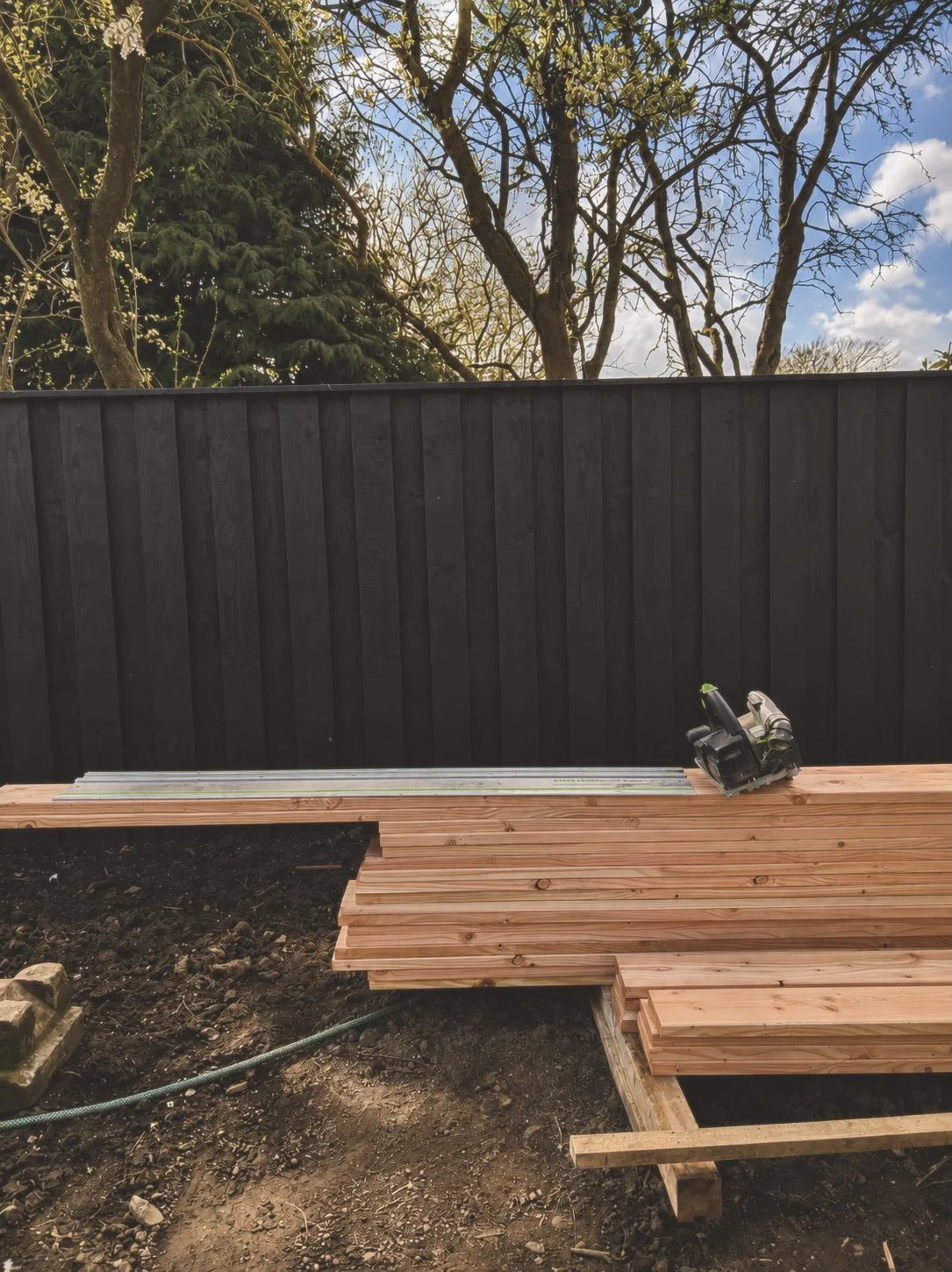 Construction site with wooden planks, a circular saw, a black fence, and a background of trees and blue sky.
