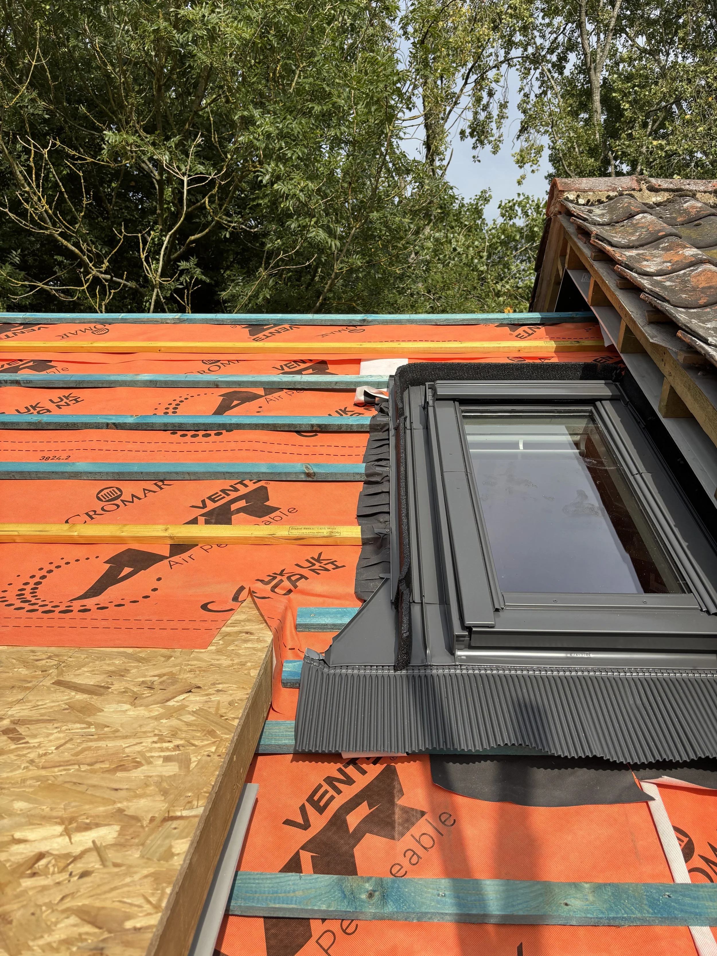 Roof under construction with insulation material, wooden beams, a roof window, and a partially completed roof structure, with trees in the background.