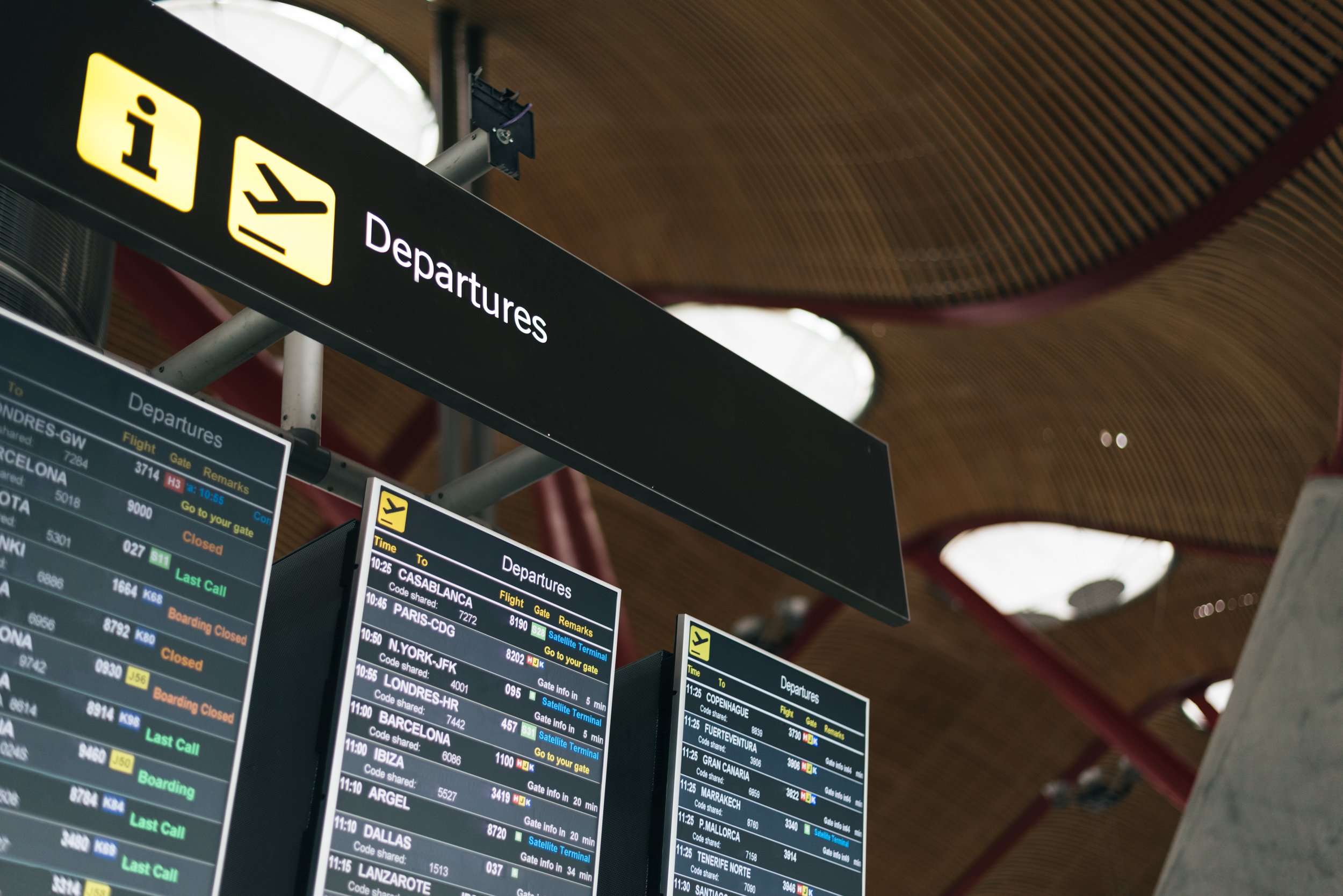 Airport departure information screens showing flight details, destinations, gate numbers, and status updates. The signs above indicate 'Departures', showing airport runs and business travel.