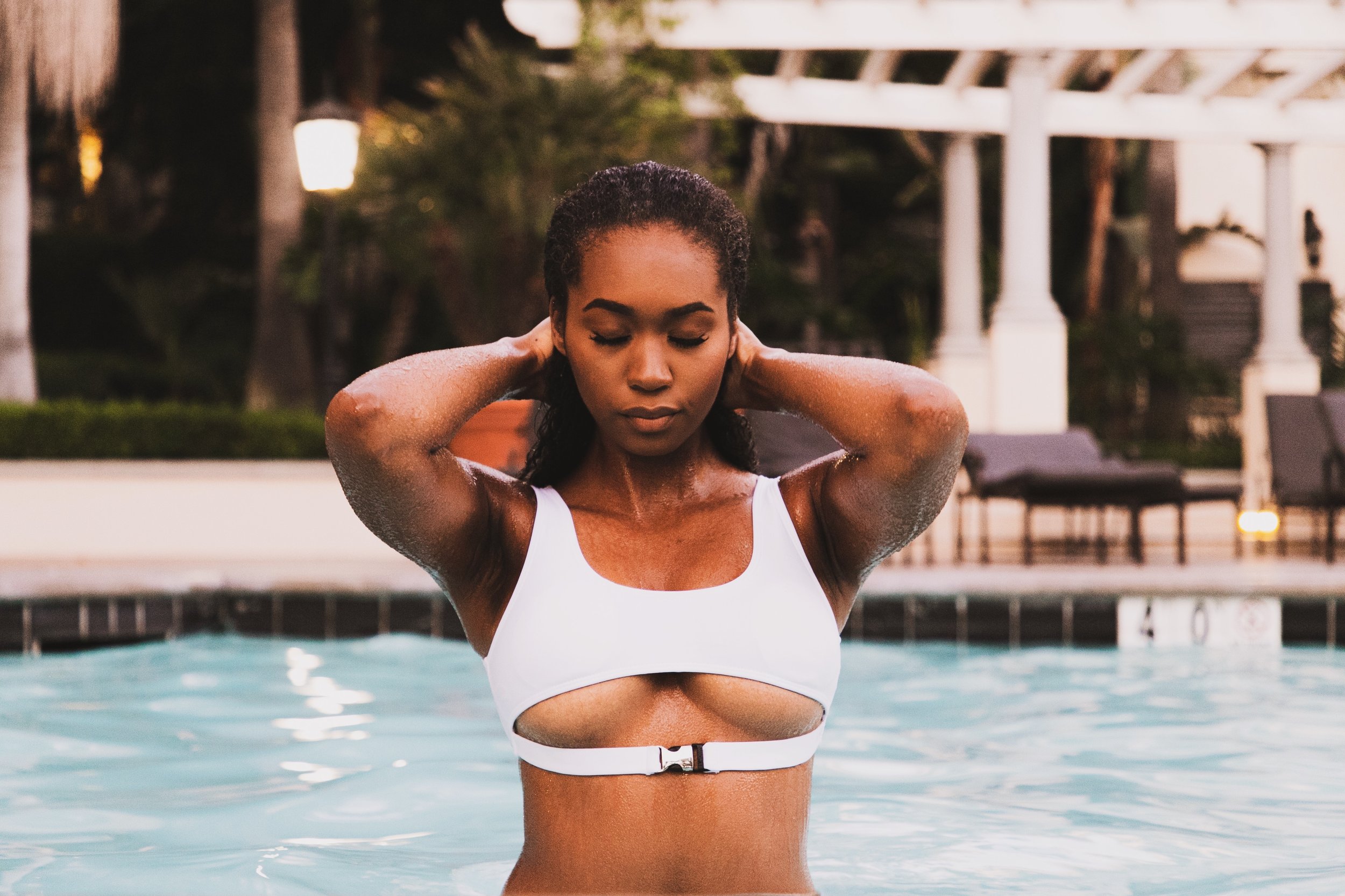 A young woman in a white swimsuit standing in a swimming pool with her hands behind her head, eyes closed, and water droplets on her arms. Background includes trees, a white structure, and outdoor lighting at dusk.