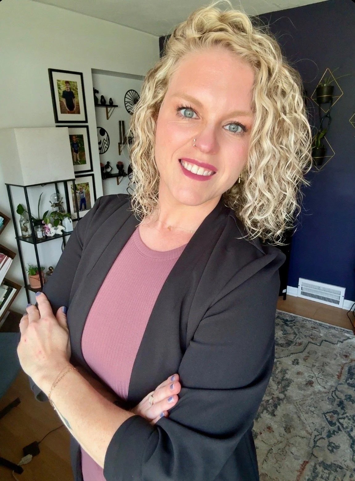 A woman with curly blonde hair and blue eyes smiling, with arms crossed, in a living room with framed photographs and shelves in the background.