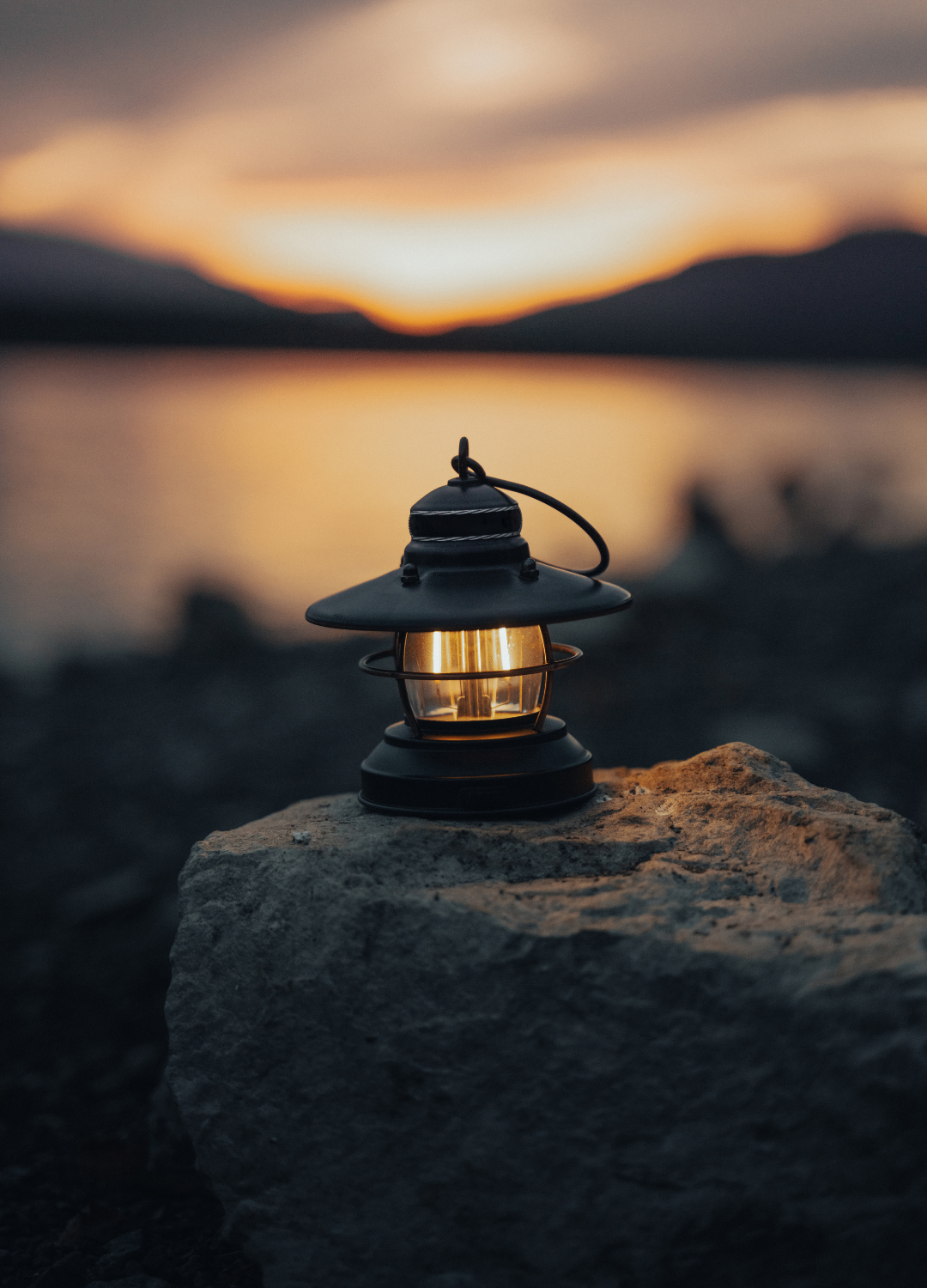 A vintage lantern resting on a rock near a body of water during sunset.