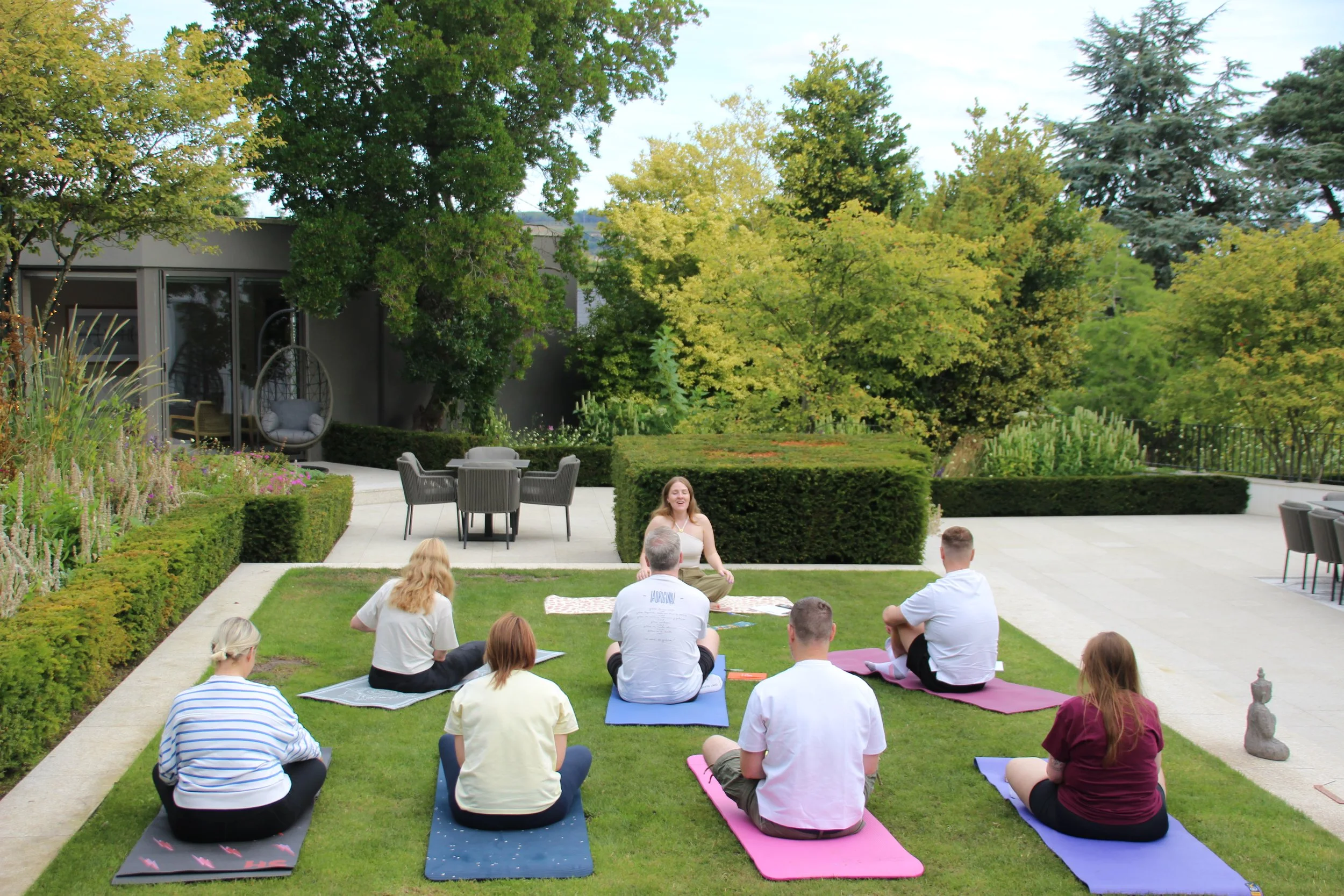 A group of people participating in an outdoor yoga or meditation session in a garden with green trees, bushes, and a modern house in the background. They are sitting on yoga mats, facing a woman leading the session.