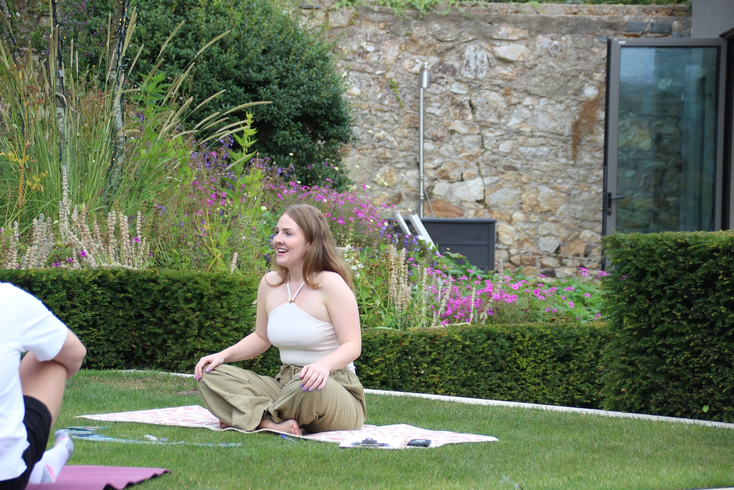 A woman with long brown hair sitting cross-legged on a patterned mat in a green garden, smiling, wearing a white tank top and khaki pants, surrounded by plants and flowers with a stone wall and glass door in the background.