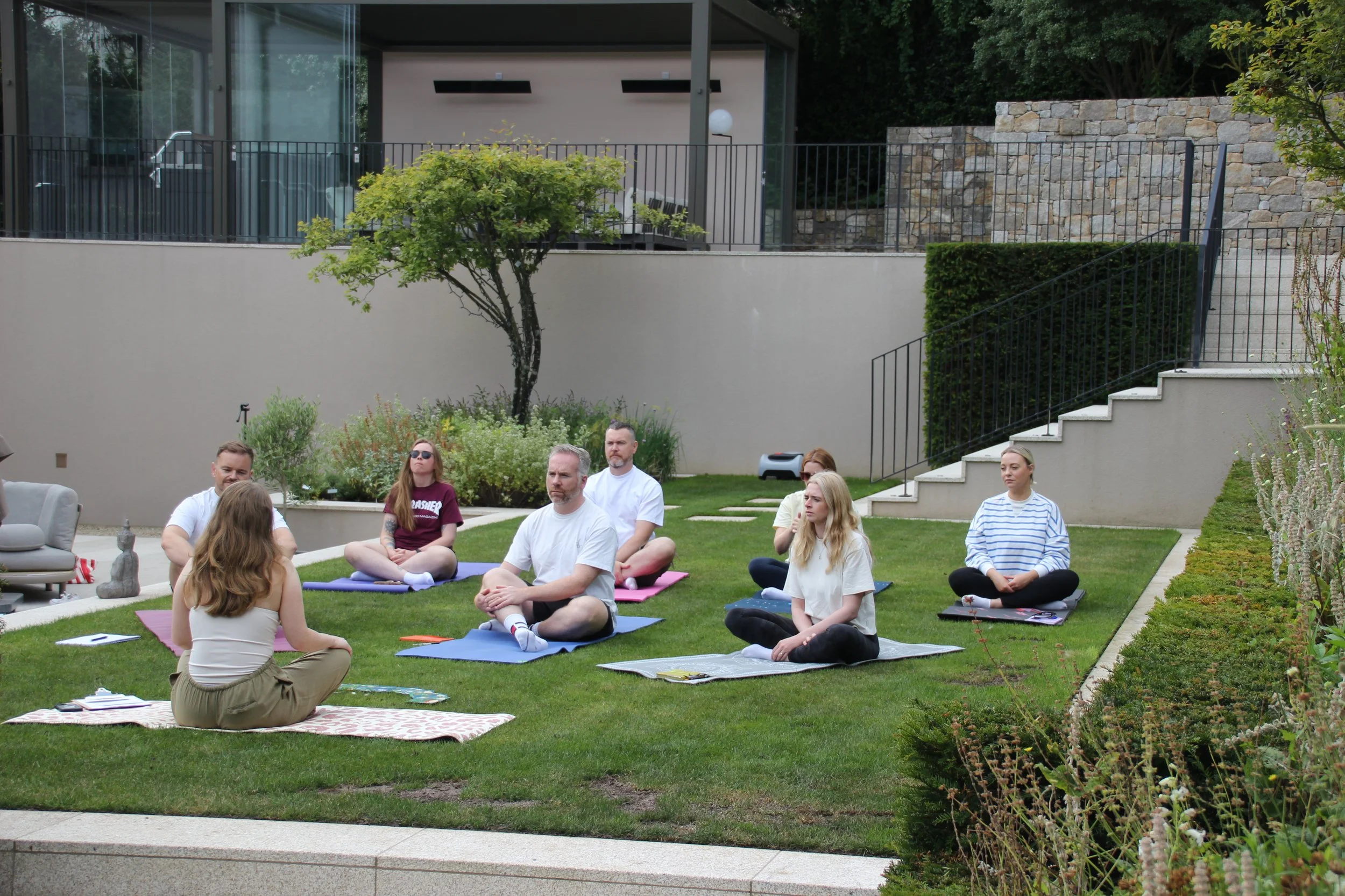 A group of people participating in an outdoor yoga or meditation class on a grassy backyard, seated on yoga mats, facing an instructor who is seated with her back to the camera. The area is landscaped with bushes, trees, and a modern house with large windows in the background.