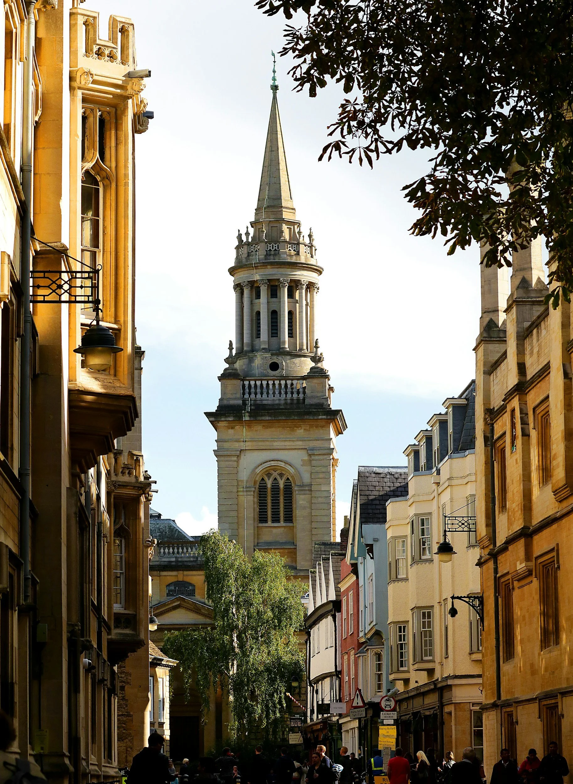 A busy street with historic buildings on both sides, a church spire in the background, and people walking below on a sunny day.