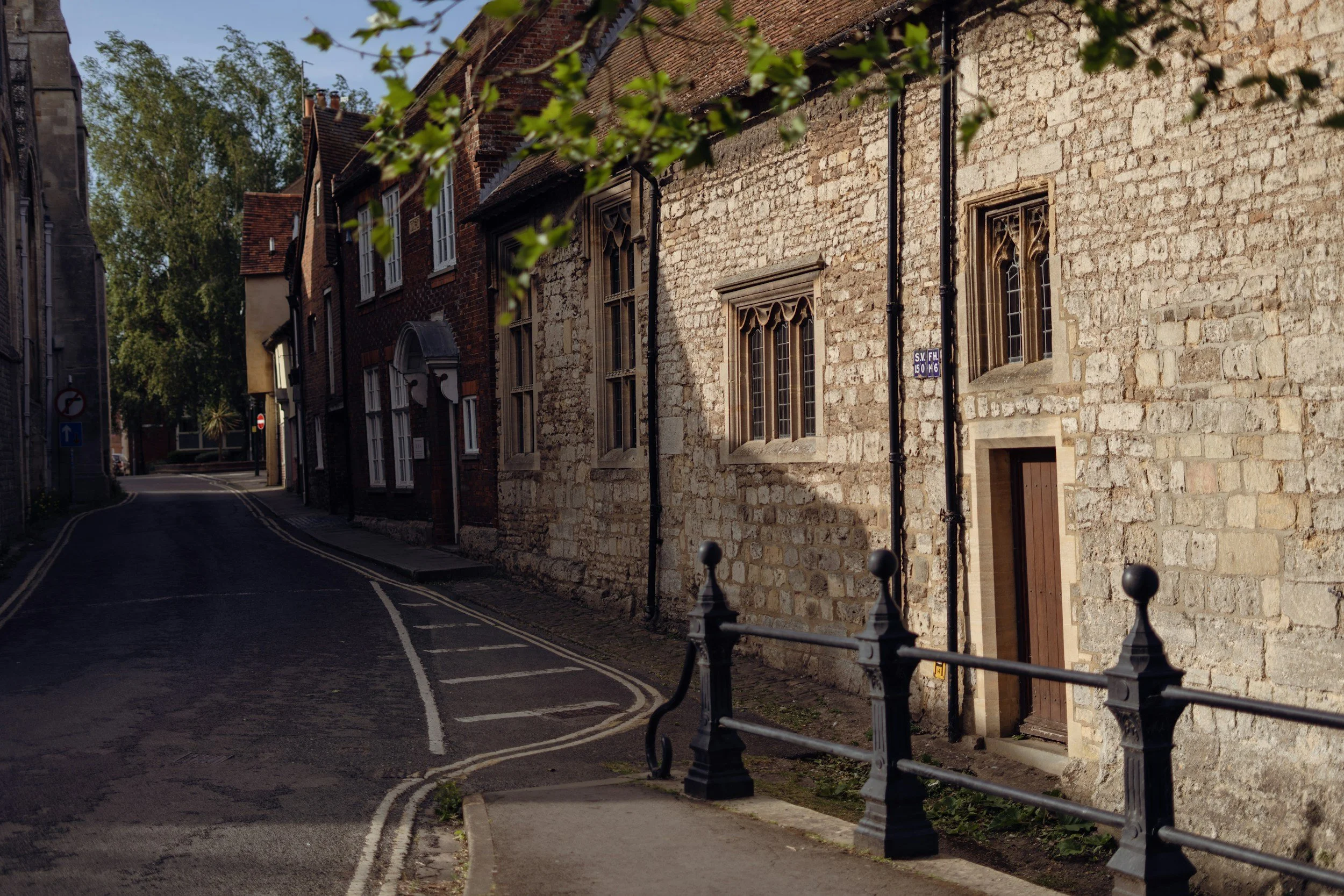 A narrow street with old brick and stone buildings on the right side and a sidewalk with black metal fencing. There are trees in the background and signs indicating traffic regulations. The scene appears to be in a historic town or neighborhood.