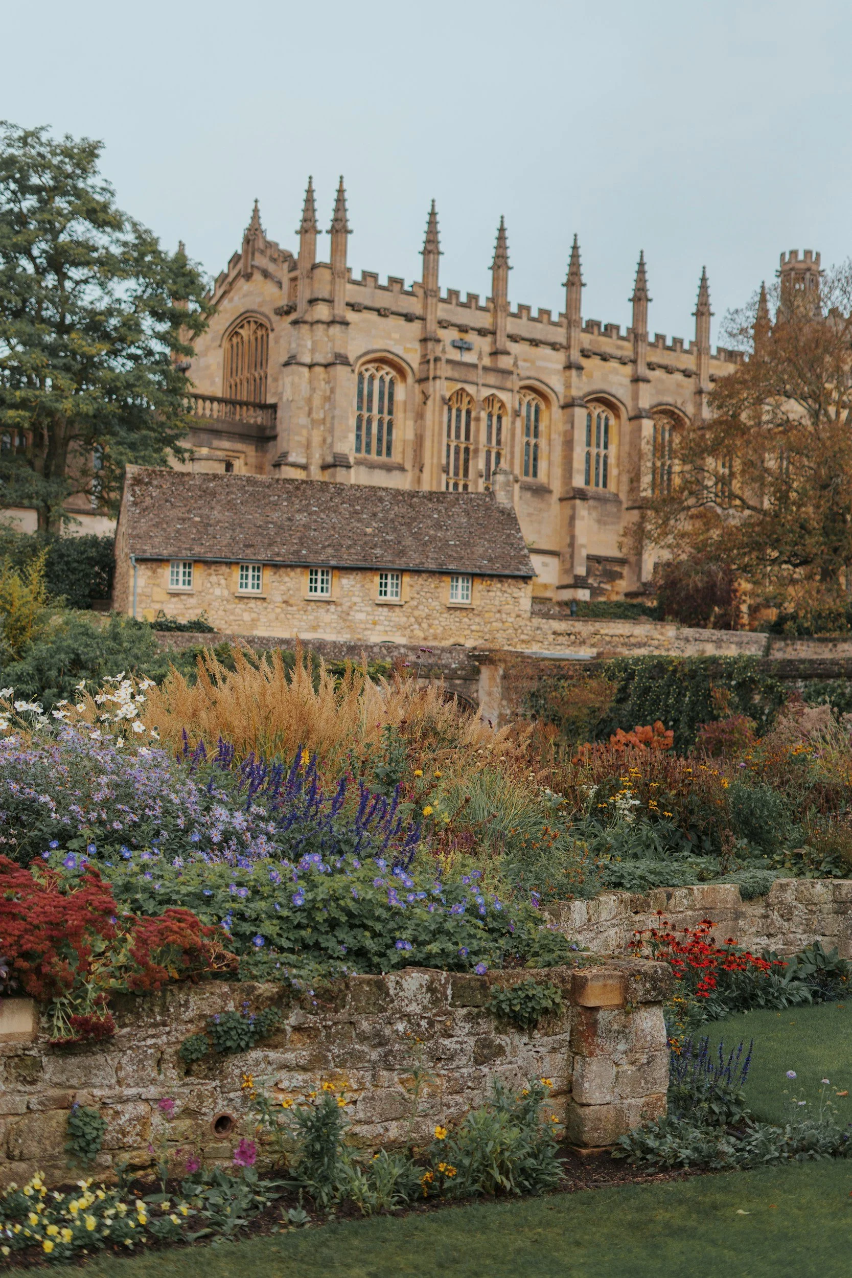 A historic stone church with gothic windows and spires, surrounded by a colorful flower garden with bushes and trees.