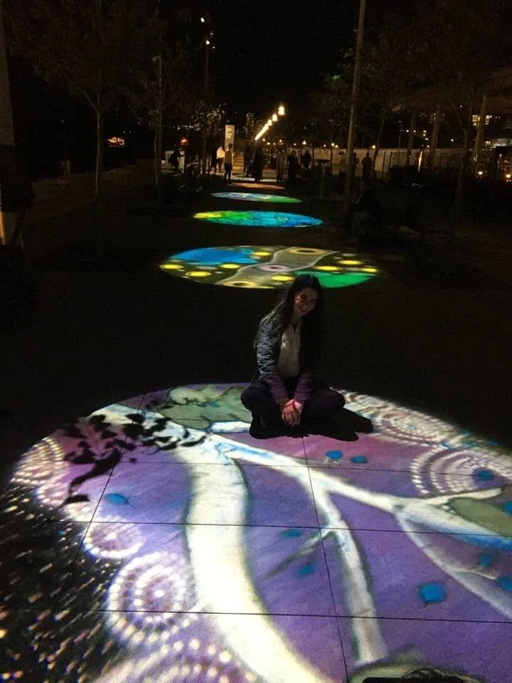 Coda Ridley sitting on her art exhibited as light projections on the ground at the Wulugul Walk at Barangaroo at Vivid Sydney.