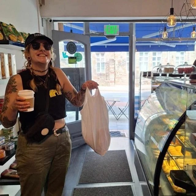 A woman with tattoos, sunglasses, and a black sleeveless shirt smiling inside a bakery or cafe, holding a coffee cup in one hand and a white grocery bag in the other, near display cases of pastries or desserts.