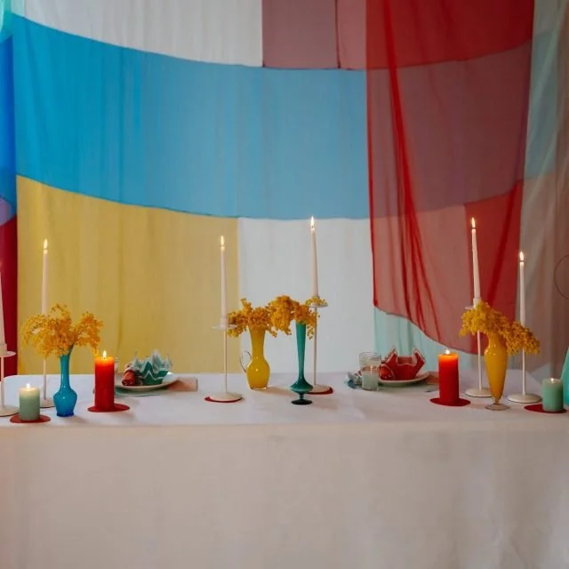 Decorative table with colorful vases holding yellow flowers, lit candles, and small bowls, with a backdrop of flowing curtains in blue, yellow, white, red, and green.