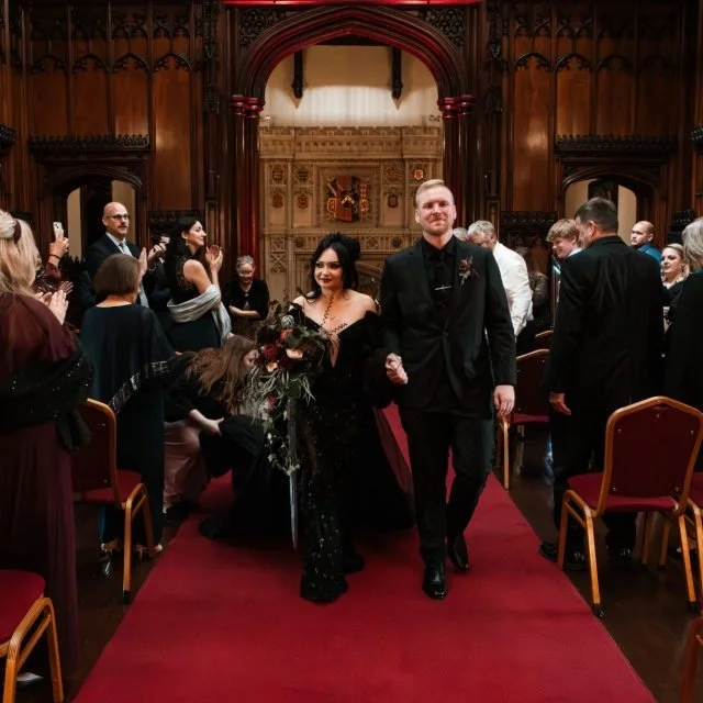A couple walking down the aisle in a formal indoor setting, celebrating an event, surrounded by guests taking photos and applauding.