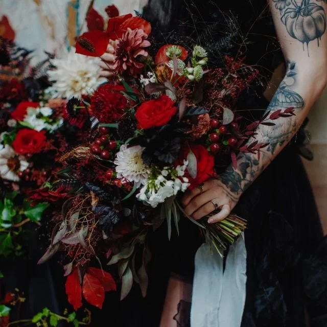 A person holding a large bouquet of red, white, and green flowers and berries, with visible tattoos on their arm.