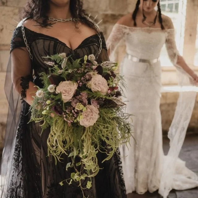 Two women in wedding dresses, one in a black gown holding a bouquet of pink and purple flowers, and another in a white dress in the background.