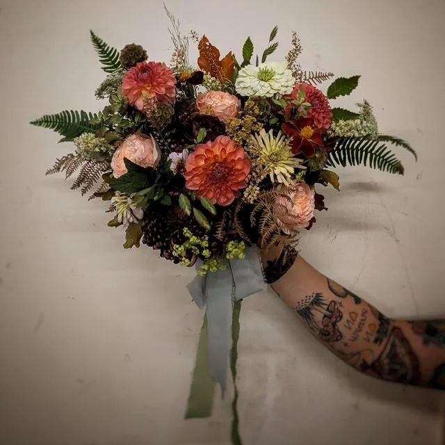 Hand holding a bouquet of mixed flowers with pink roses, orange dahlias, white daisies, and various green leaves against a plain background.