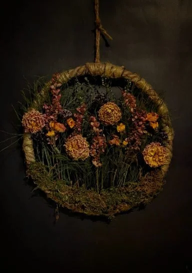 A circular floral arrangement with orange and purple flowers hanging from a brown rope against a dark background.