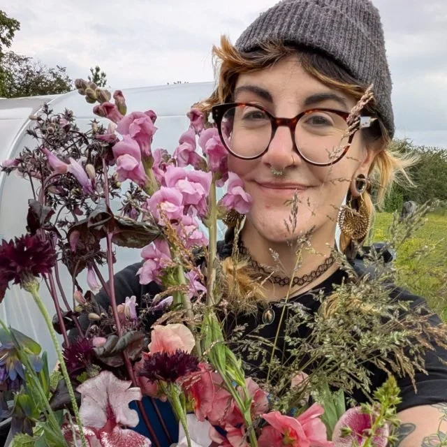 Person smiling, holding a bouquet of pink and purple flowers outdoors, with trees and a cloudy sky in the background.