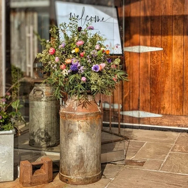 Vintage metal milk cans filled with fresh flowers positioned next to a rustic wooden wall and a glass Welcome sign.