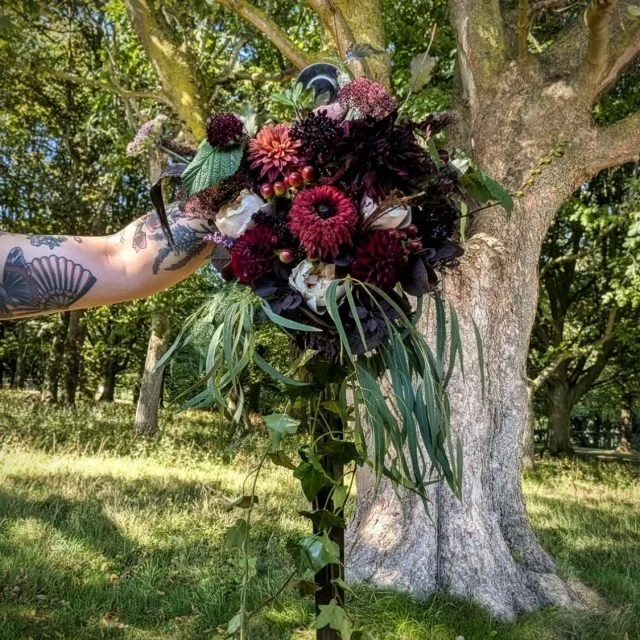 Person holding a large bouquet of dark and light red flowers with greenery, in front of a tree in a park.