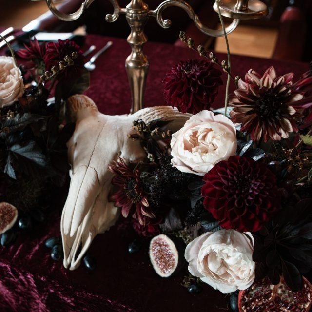 Decorative arrangement featuring a deer skull with antlers, surrounded by dark red, white, and purple flowers, and black berries, on a table with a dark red tablecloth.