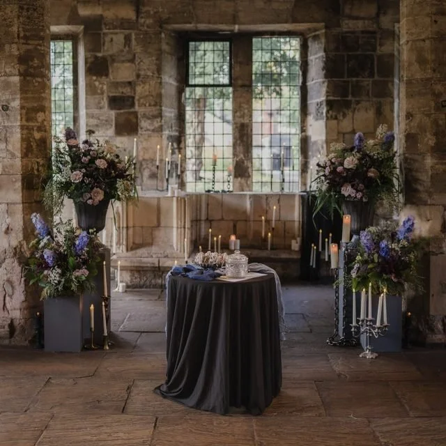 A decorated table with a black tablecloth, candles, and a cake in front of a stained glass window in a stone wall room with floral arrangements and candles.