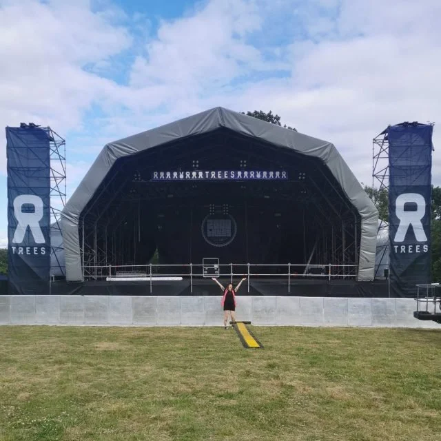 Empty outdoor stage with large black canopy, two flag-like banners on each side with a white 'R' and 'TREES', and a person standing in front with arms raised, on a grassy field.