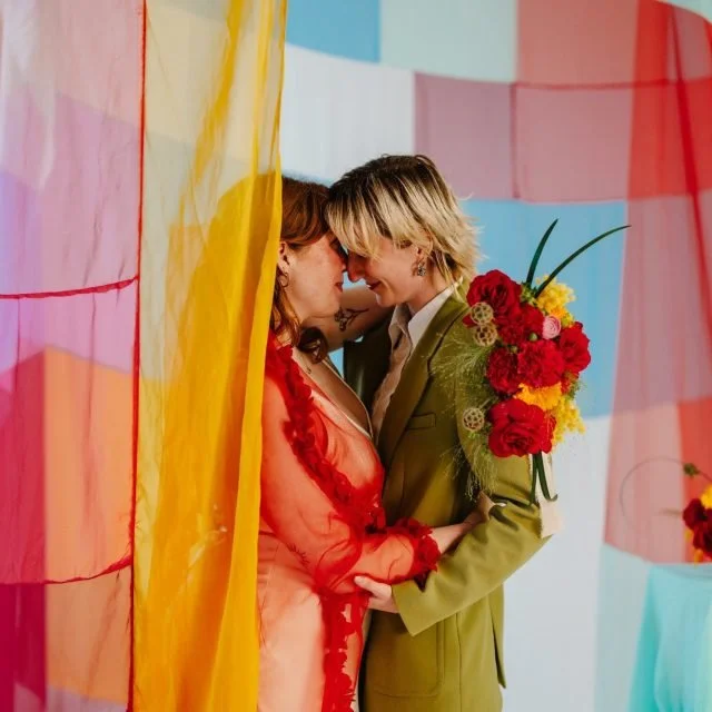 A couple stands close, touching foreheads and smiling, behind colorful fabrics, with the woman holding a bouquet of red, yellow, and pink flowers.