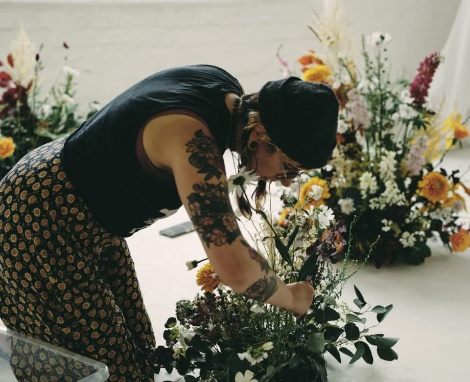 A woman with tattoos, glasses, a black beanie, wearing a black sleeveless top and patterned pants, arranging a floral bouquet with various flowers and greenery.