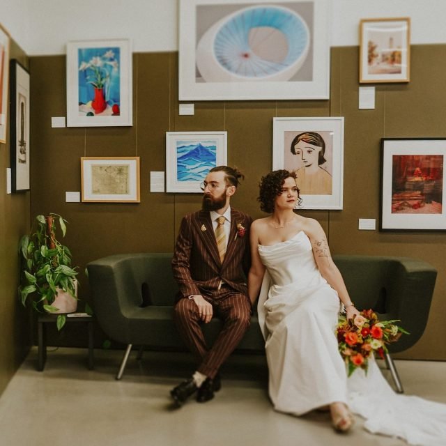 A bride and groom sitting on a green sofa in an art gallery, with the bride holding a bouquet of flowers. The groom is dressed in a brown pinstripe suit, and the bride is wearing a white strapless wedding gown. Artworks are displayed on the wall behi