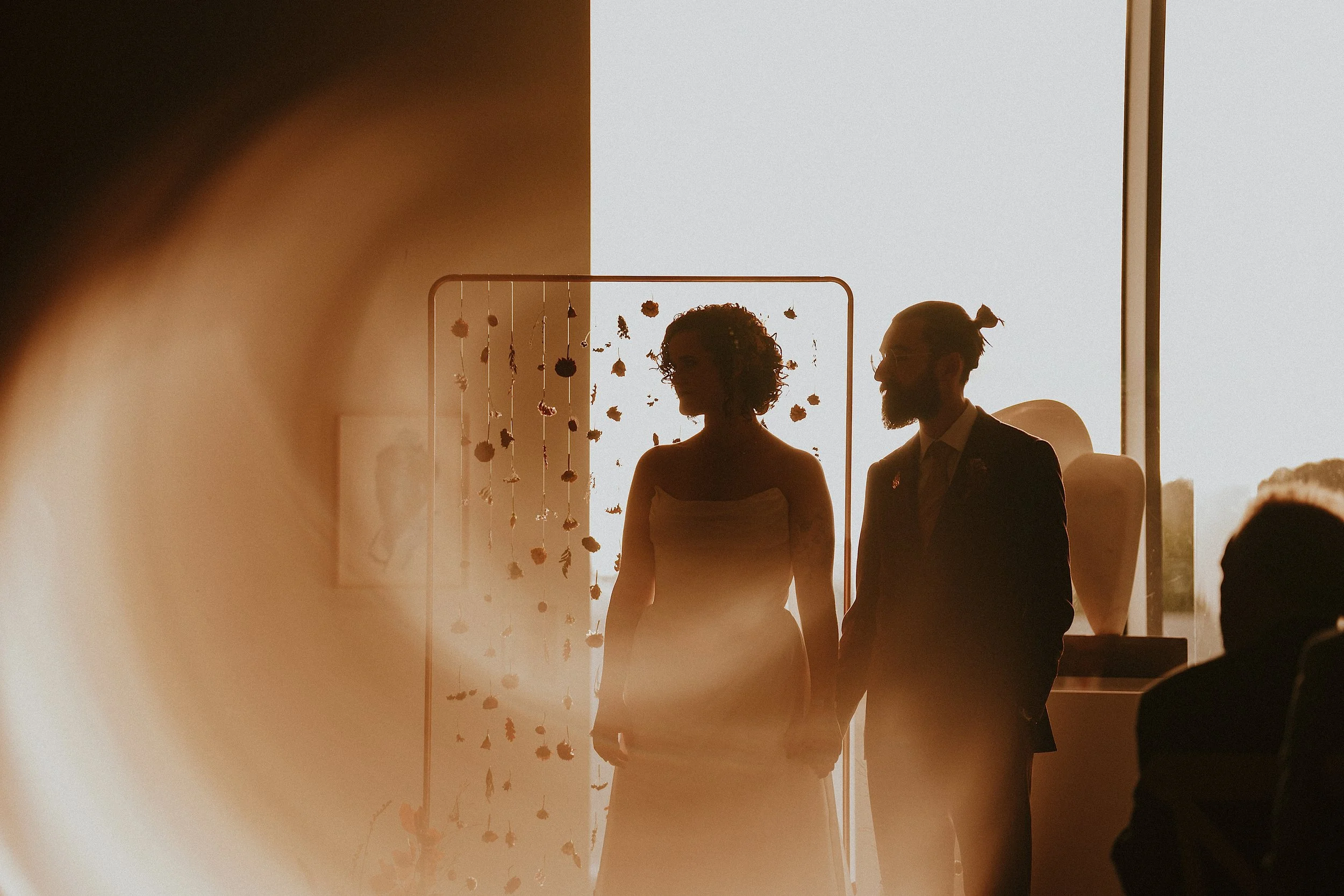 Silhouettes of a bride and groom standing beside each other during a wedding ceremony indoors with a bright window in the background and floral decorations hanging.