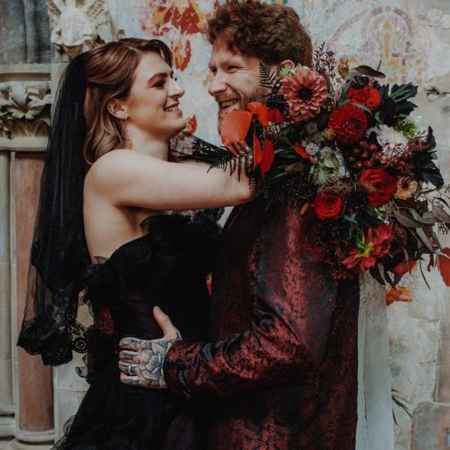 A couple smiling and looking into each other's eyes, with the woman embracing the man and holding a large bouquet of flowers.