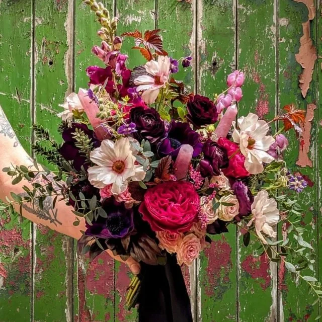 A large bouquet of mixed flowers, including roses, anemones, and other blooms, held in someone's arm against a weathered green wooden background.