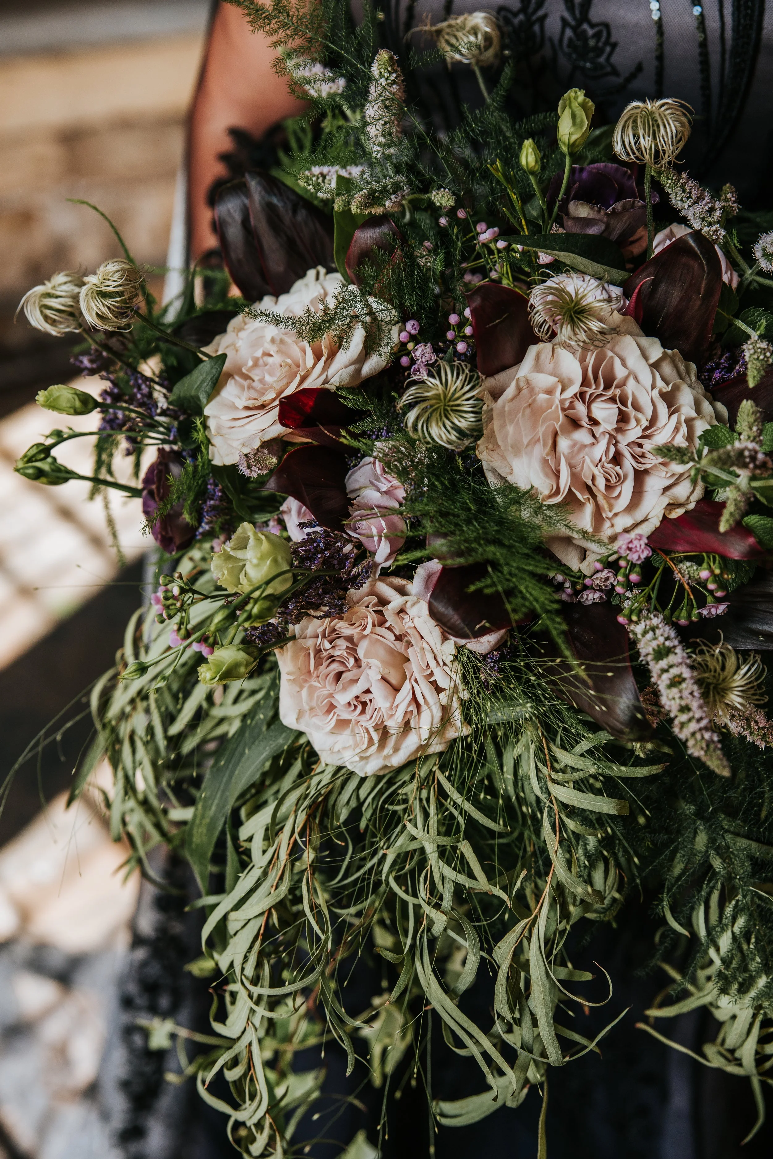Close-up of a lush bouquet of flowers with light pink roses, dark purple calla lilies, white and purple veronica, green foliage, and various other blooms.