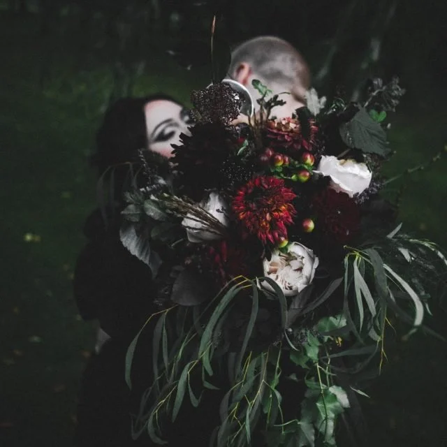 A person with dark hair and makeup holding a large dark floral bouquet covering their face in a lush green outdoor setting at night